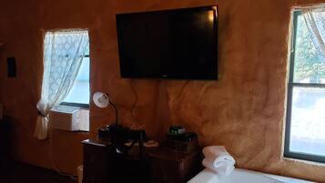 Quarto com televisão de tela plana na parede, uma escrivaninha com abajur, relógio e janela com cortinas estampadas.
