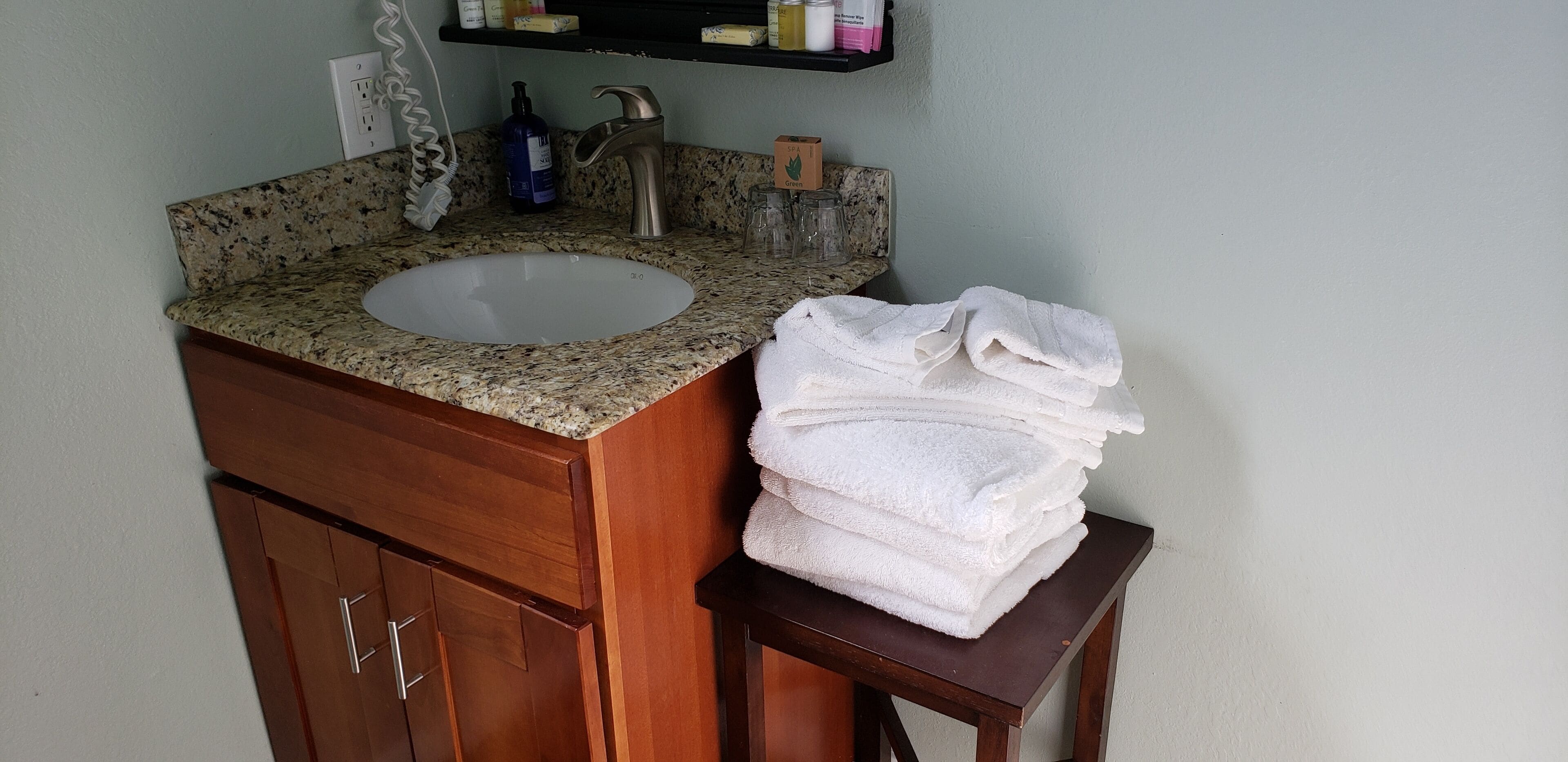 A bathroom with a granite countertop sink, a wooden cabinet below, and a stack of white towels on a small table.