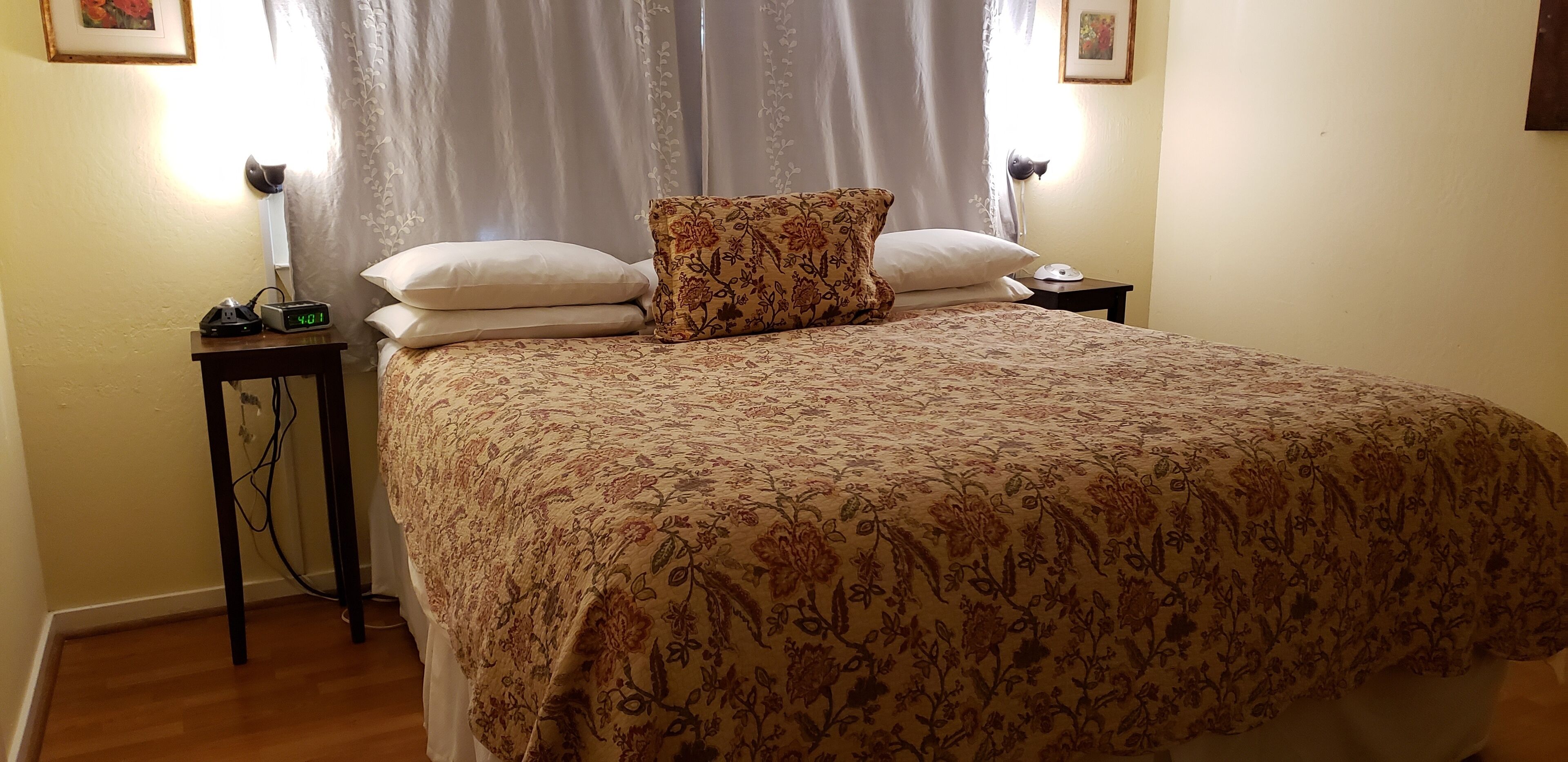 Een netjes opgemaakt bed met een bloemenprintdeken en twee kussens, een nachtkastje met een lamp en een klok, en een gordijn dat deels is opengetrokken.