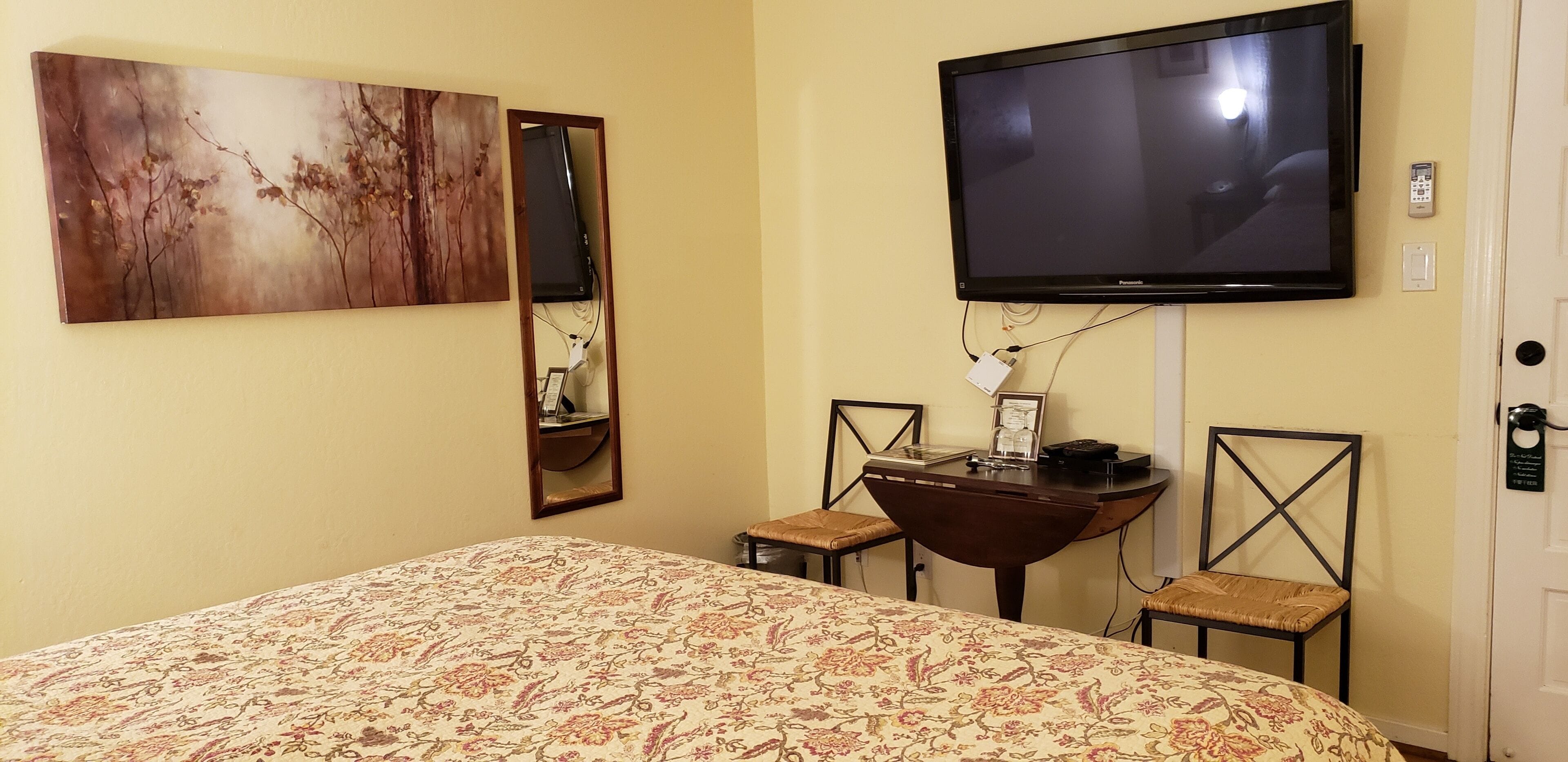Une chambre d’hôtel avec un grand lit, un téléviseur à écran plat fixé au mur, une petite table en bois avec des chaises et un grand miroir.