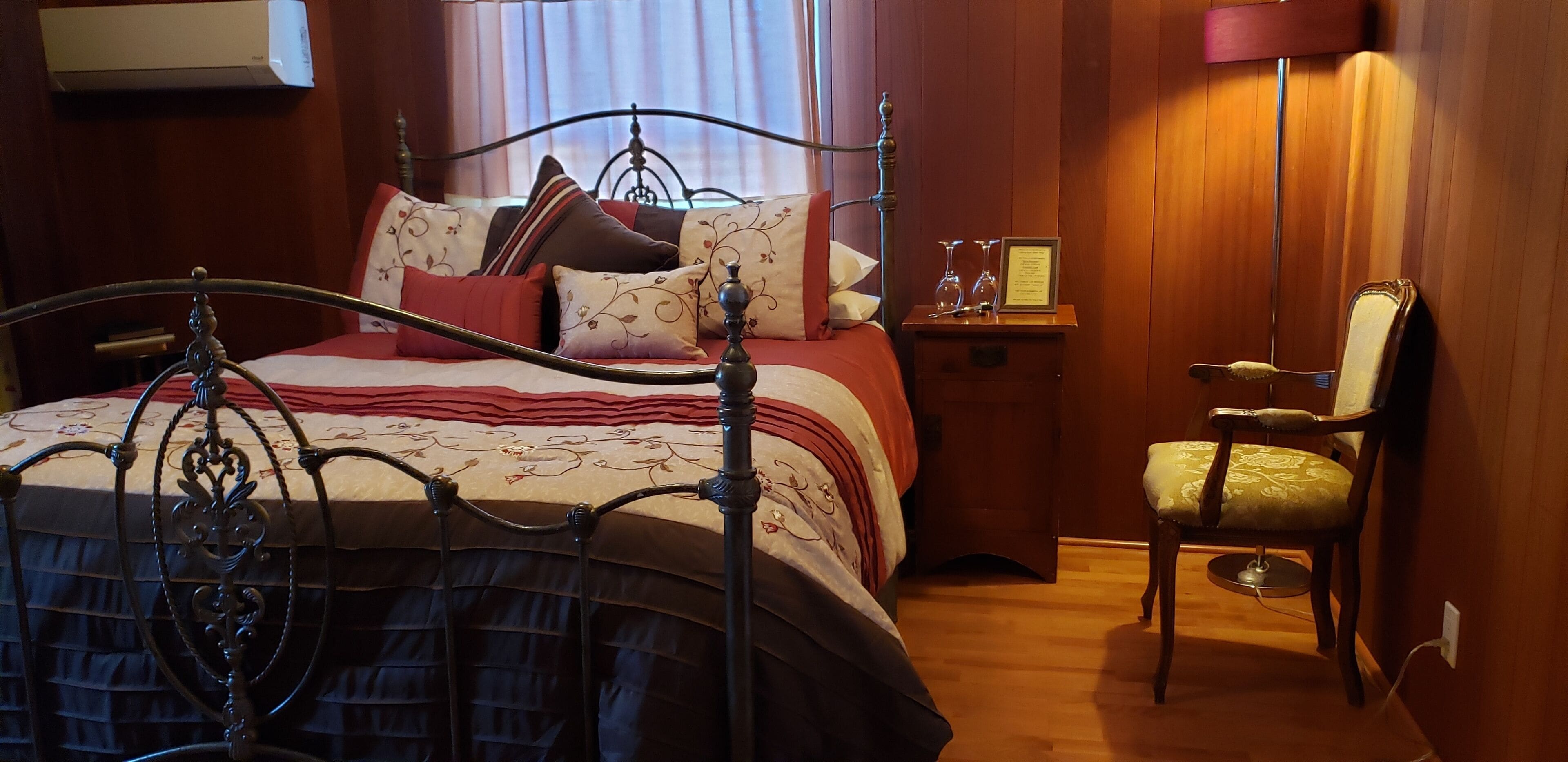 A neatly made bed with a metal frame, a bedside table with a lamp, and a chair with a cushion.