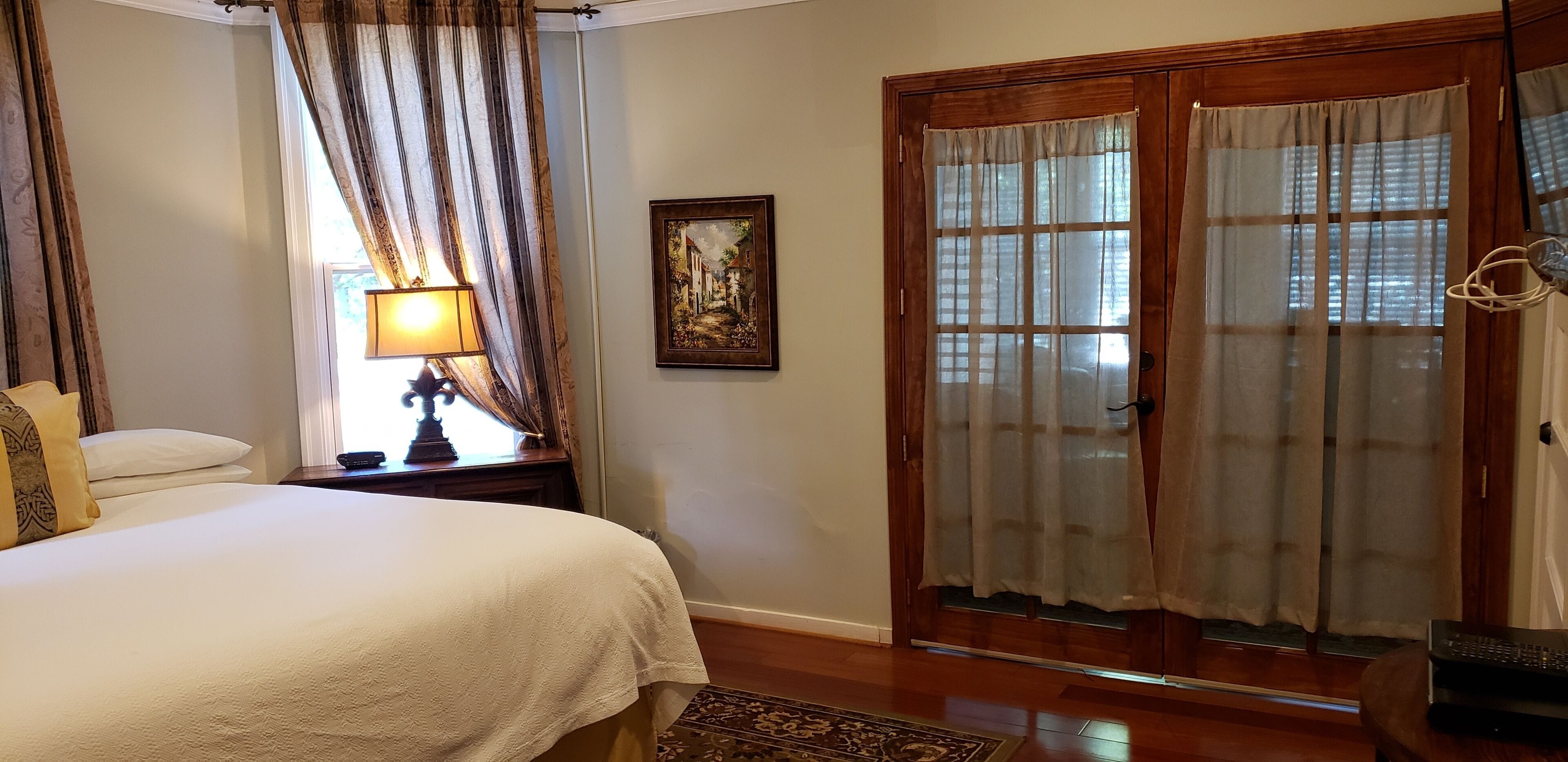 Un dormitorio con cama, mesita de noche con lámpara, un cuadro y puerta corrediza de vidrio.