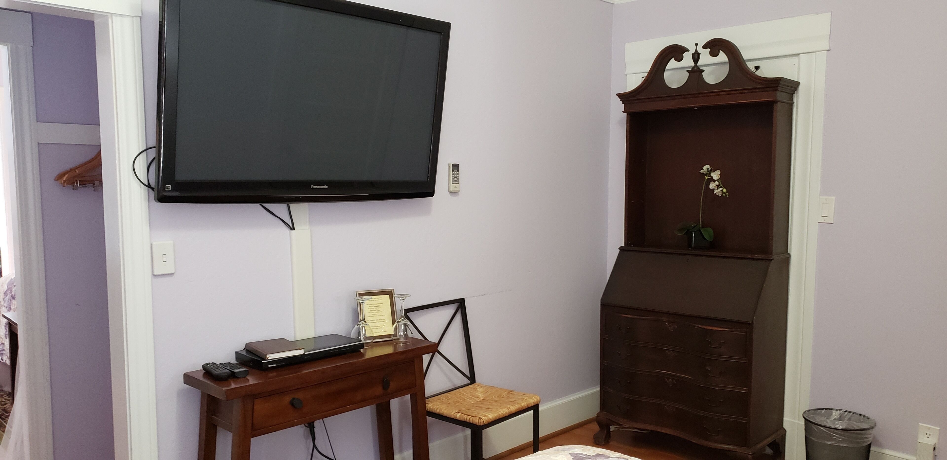Een kamer met een wandtelevisie, een houten dressoir, een klein bureau met stoel en een prullenbak.