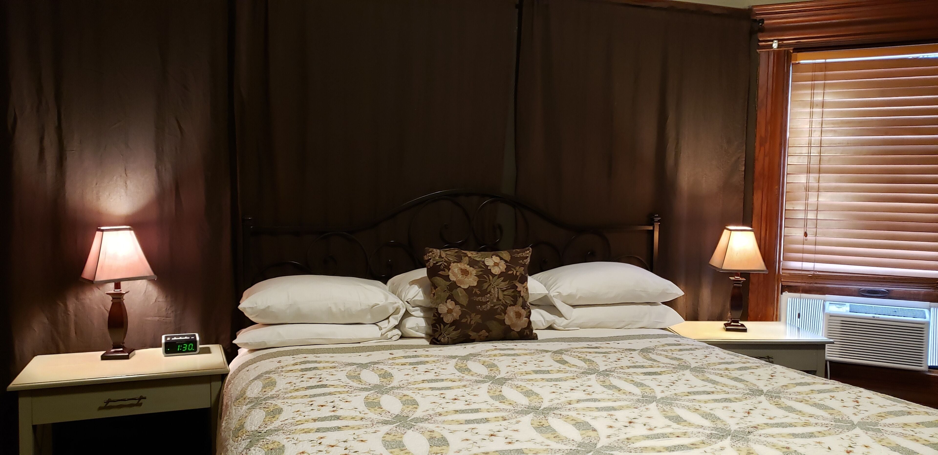 Una cama bien hecha con colcha estampada con motivos florales, dos lámparas de noche, un reloj digital y una ventana con persianas.