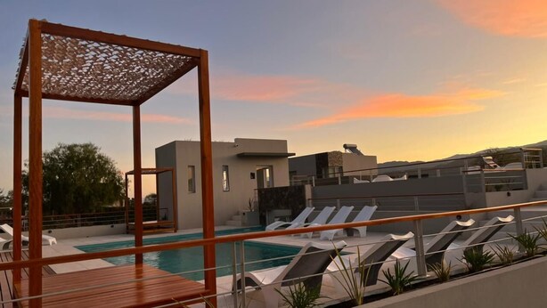 Un área de piscina en la azotea con sillones de descanso, una pérgola de madera y un edificio moderno al fondo.