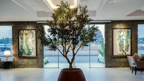 A lobby with a large potted tree, framed artwork on the walls, and a view of a cityscape through large windows. - Viola Gardens Residence ( economic serviced apartments) (Riyadh)