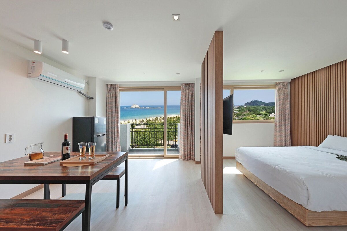 A hotel room with a bed, a desk with a TV, a minibar, and a balcony with a view of the sea.