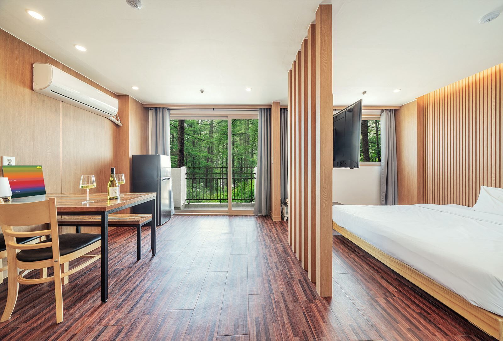 A modern hotel room with a wooden floor, a bed, a desk with a chair, a TV, and a balcony with a view of trees.