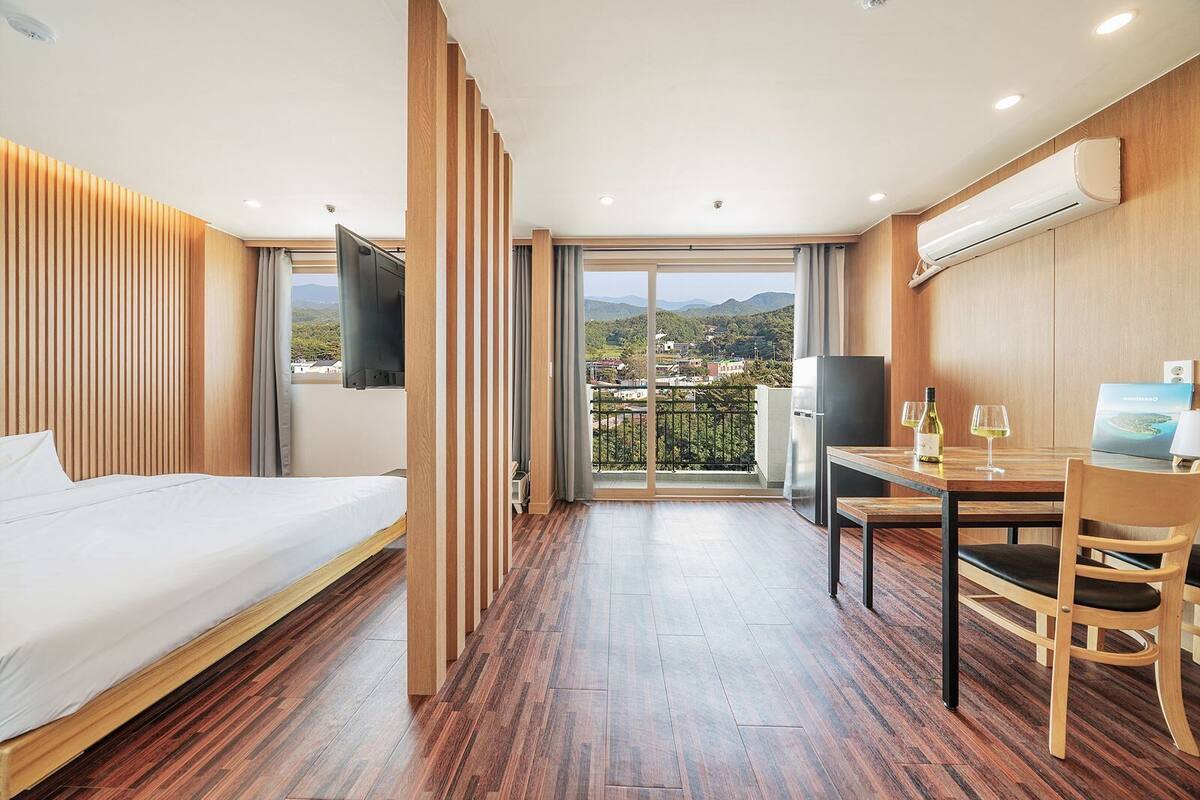 A hotel room with a bed, a dining table with chairs, a TV mounted on the wall, and a view of the landscape through the window.
