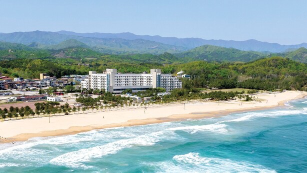 Un hotel costero con varias edificaciones, una playa de arena y vistas a colinas verdes al fondo.