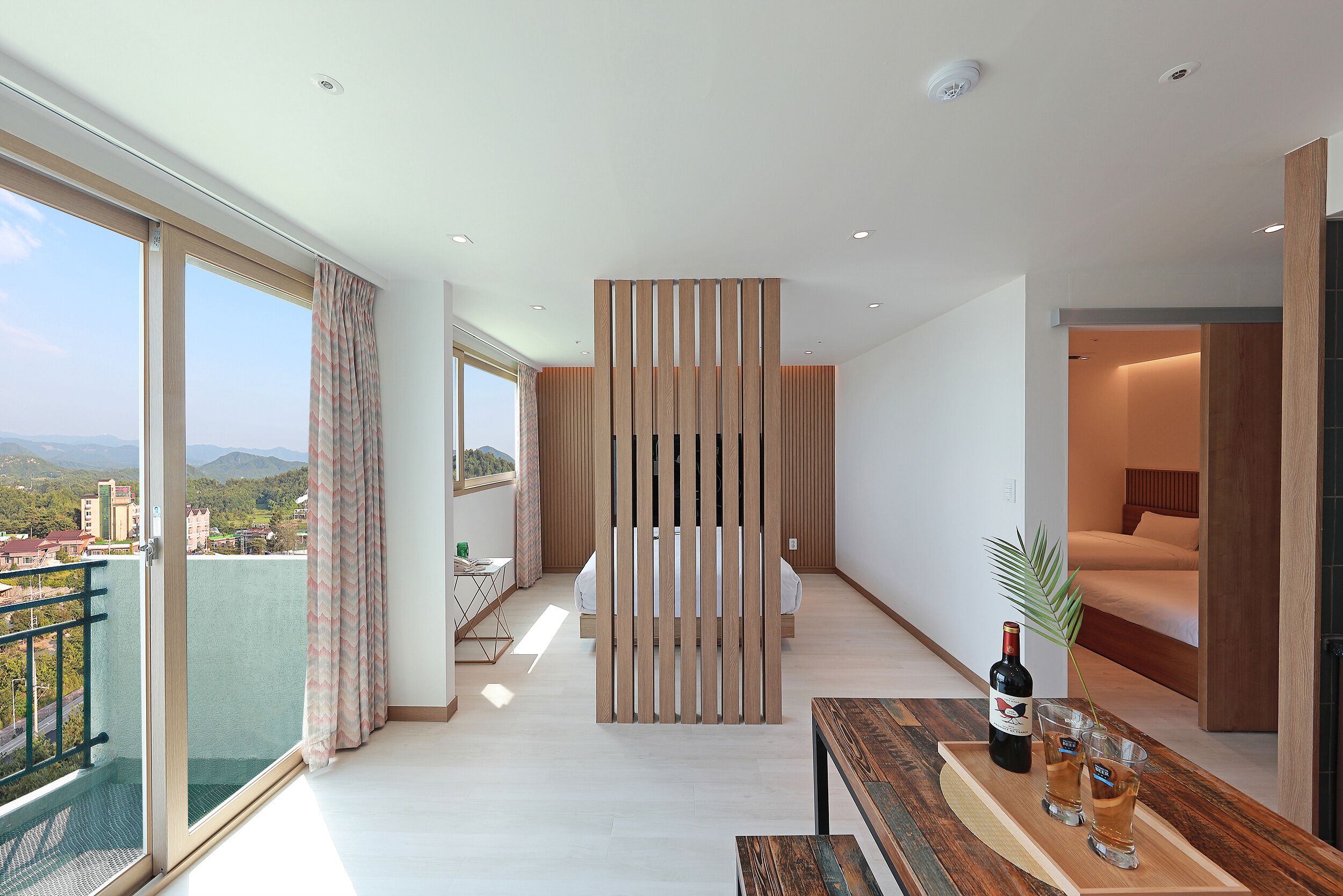 A modern hotel room with a balcony, a bed, a wooden partition, and a dining area with a bottle and glasses.