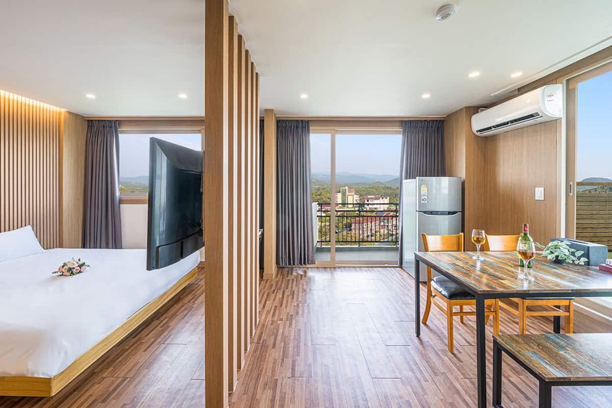 A modern hotel room with a bed, a TV, a dining table, and a balcony with a view of the countryside.