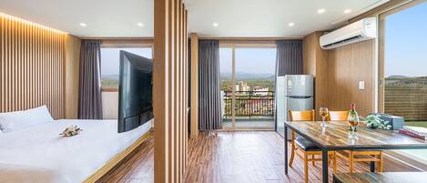 A modern hotel room with a bed, a TV, a dining table, and a balcony with a view of the countryside.