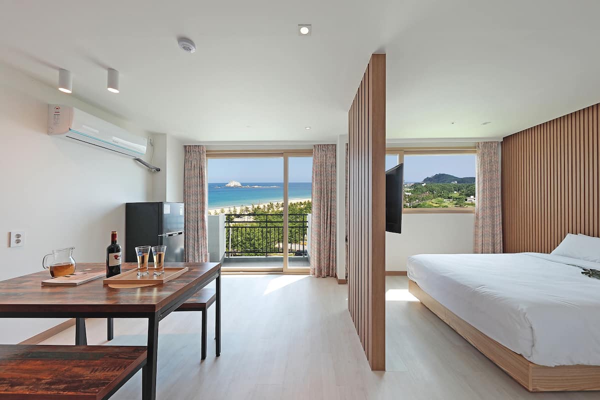 A modern hotel room with a bed, a desk with a TV, a balcony with a view of the sea, and an air conditioning unit.