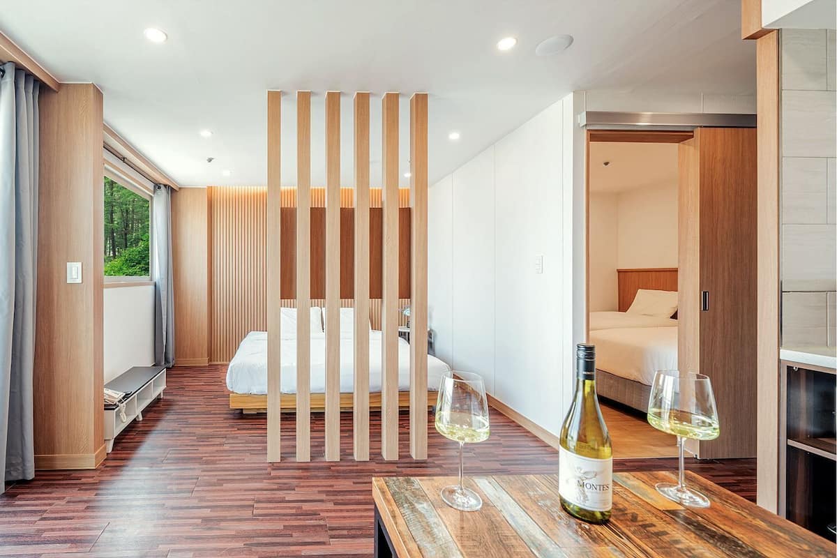 A modern hotel room with a wooden floor, a bed with white linens, a dining area with a wooden table and wine glasses, and a view of the forest through large windows.