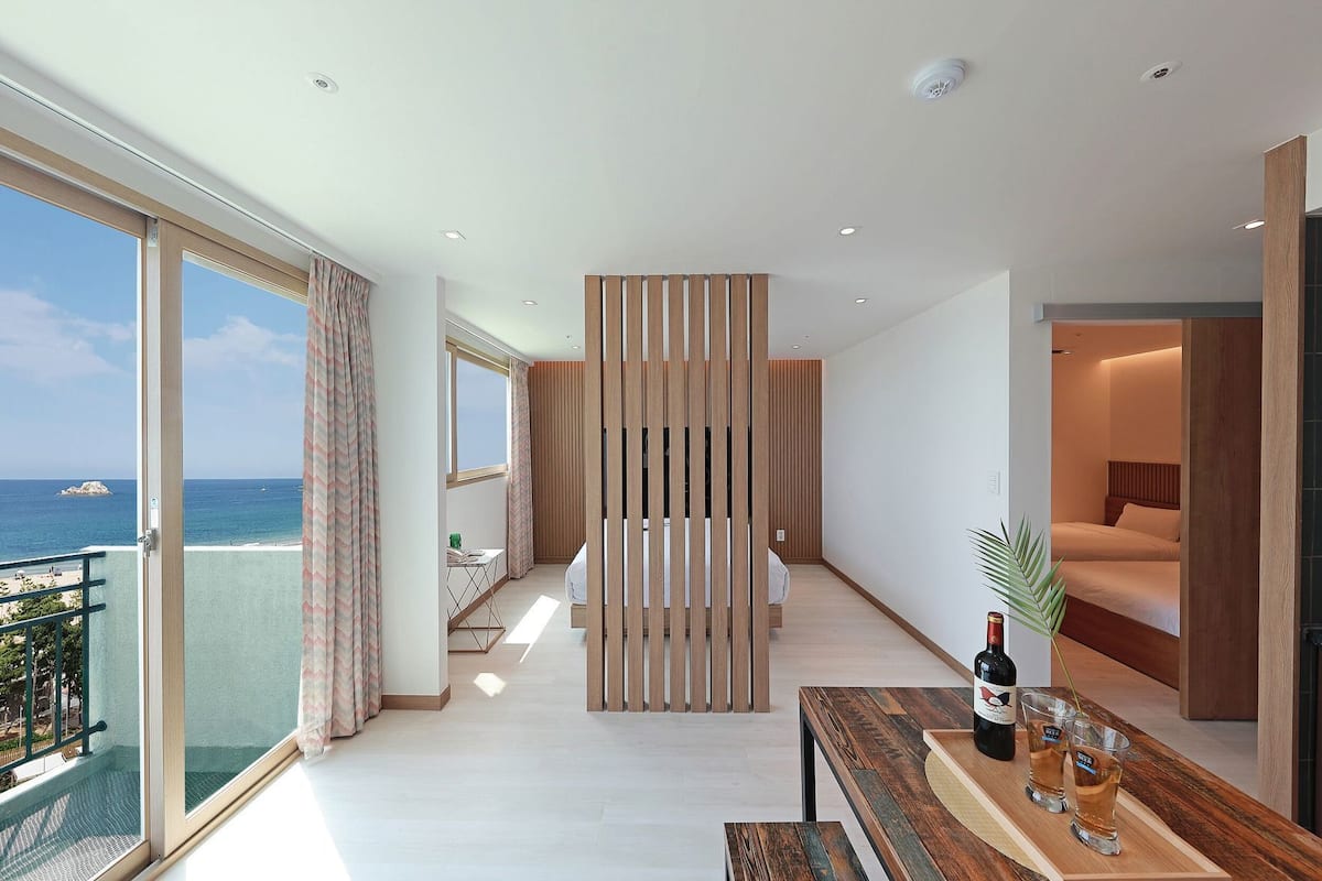 A modern hotel room with a balcony, a wooden partition, a bed, a desk with a bottle and glasses, and a view of the sea.