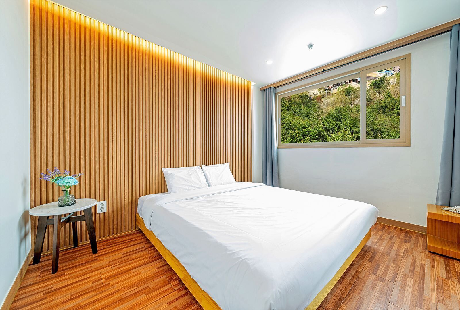 A modern bedroom with a wooden slat wall, a large bed, a small round table with a vase, and a window with a view of greenery.