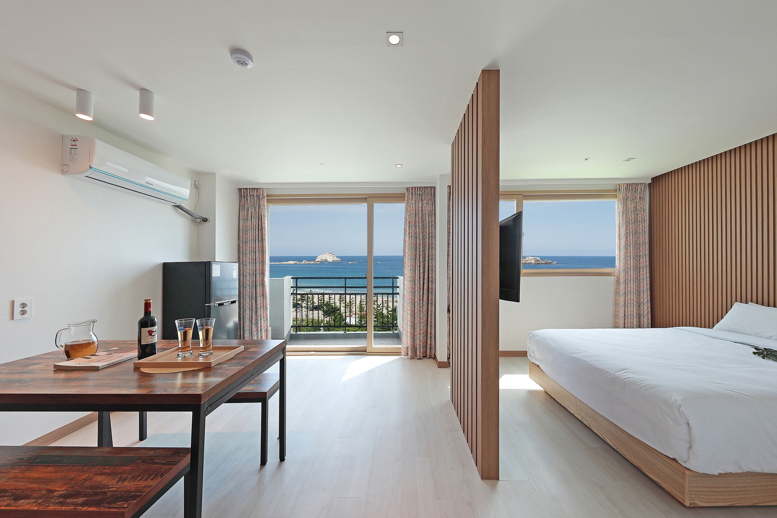A modern hotel room with a bed, a desk with a TV, a balcony with a view, and an air conditioning unit.