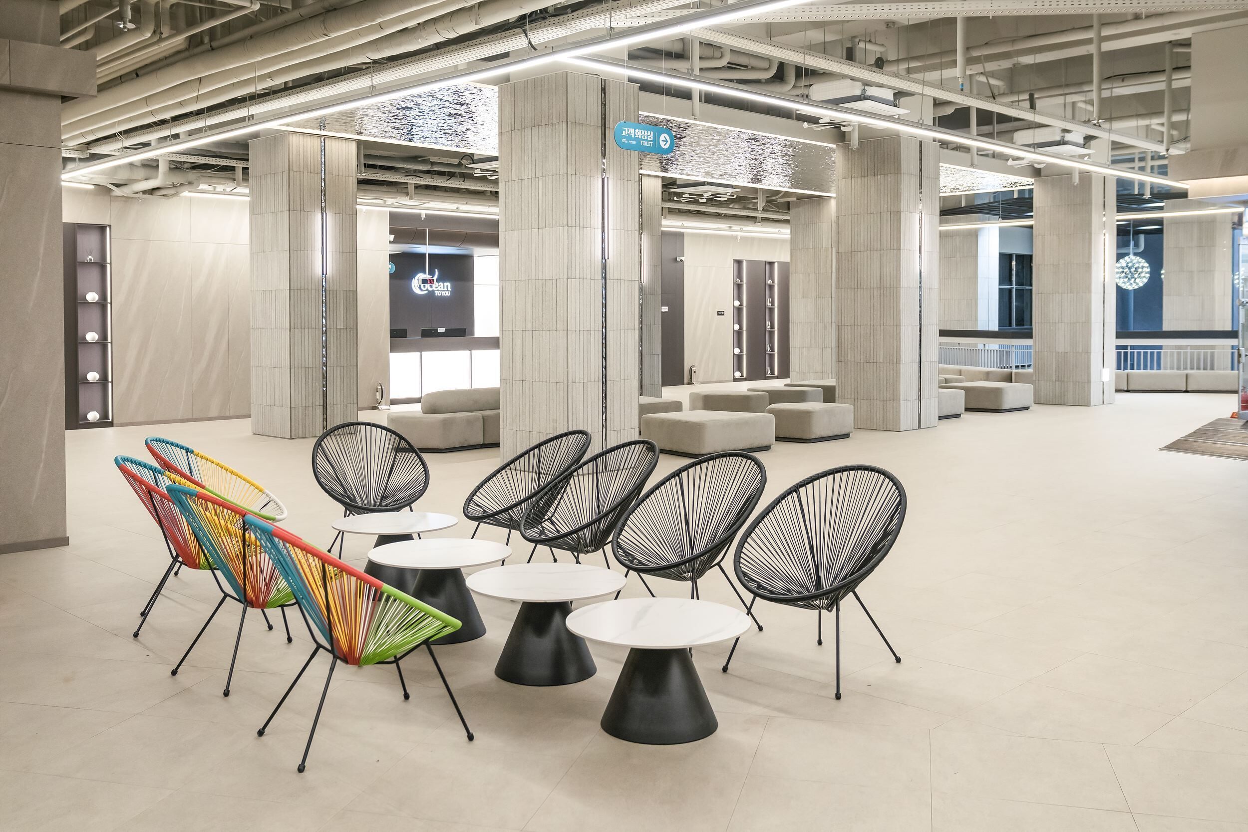 A modern indoor space with high ceilings, exposed pipes, and a mix of seating arrangements including colorful chairs and black wireframe chairs around small round tables.