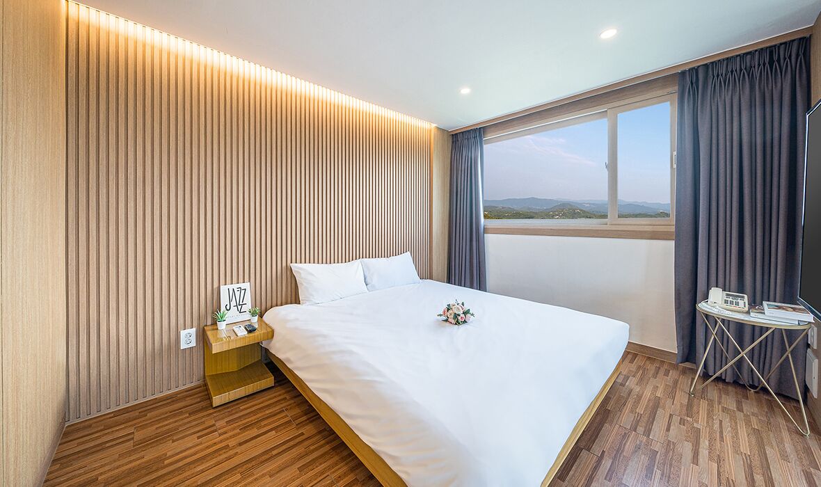 A modern hotel room with a large bed, a bedside table with a lamp, a small table with a phone and a book, and a view of the outdoors through a large window.