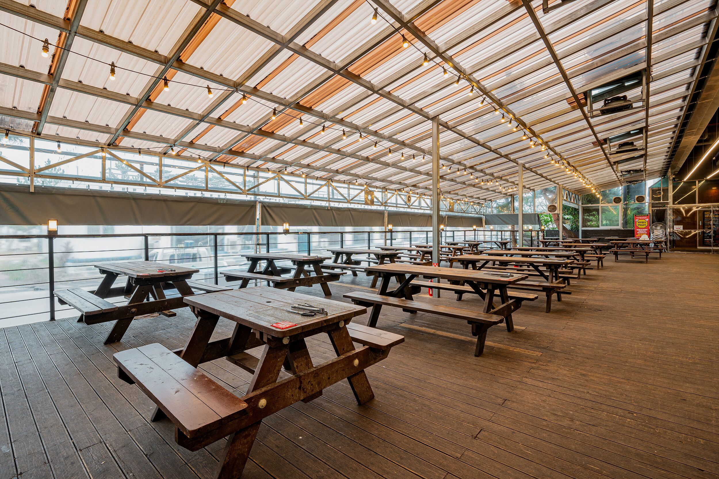An outdoor covered picnic area with wooden tables and benches, large windows, and a view of the water.