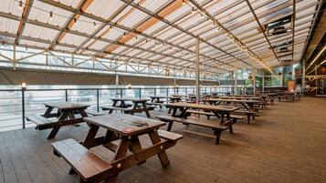 An outdoor covered picnic area with wooden tables and benches, large windows, and a view of the water.