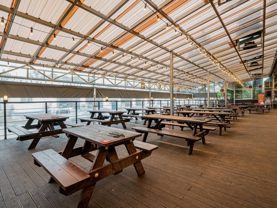 An outdoor covered picnic area with wooden tables and benches, large windows, and a view of the water.