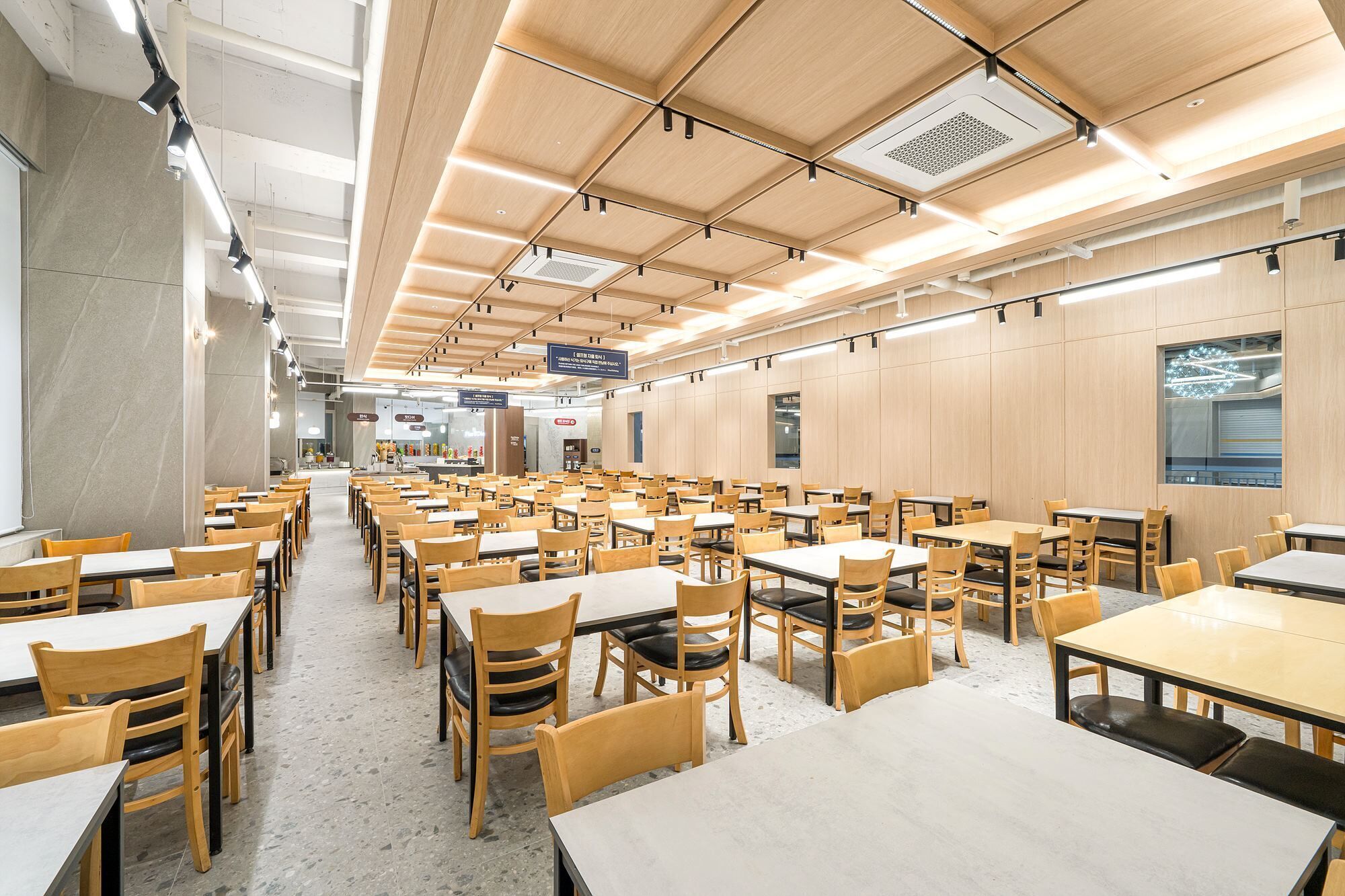 A spacious cafeteria with multiple tables and chairs, high ceilings with exposed beams, and a clean, modern design.