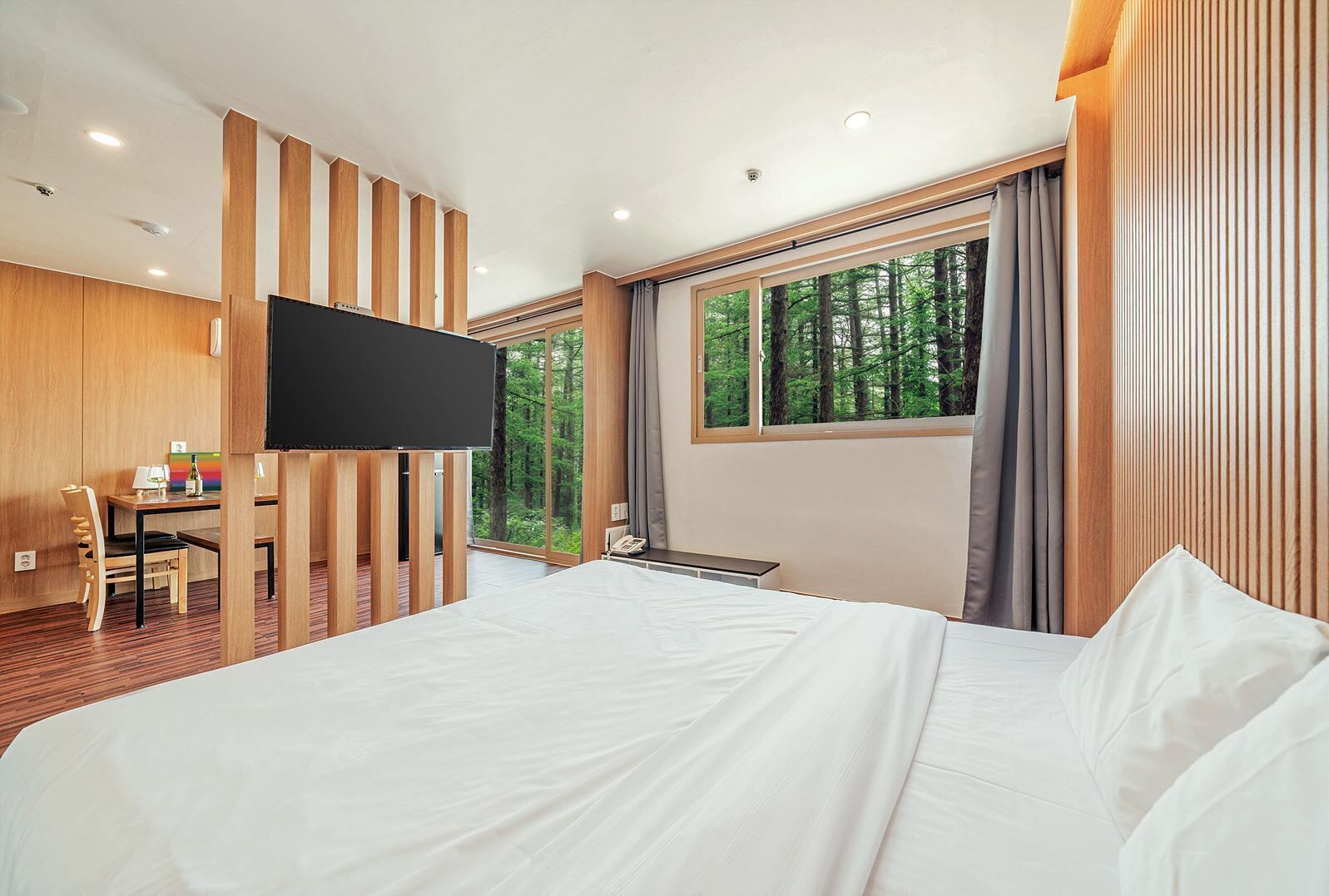 Une chambre d’hôtel moderne dotée d’un grand lit, d’un bureau avec une chaise, d’une télévision à écran plat et d’une grande fenêtre donnant sur la forêt.
