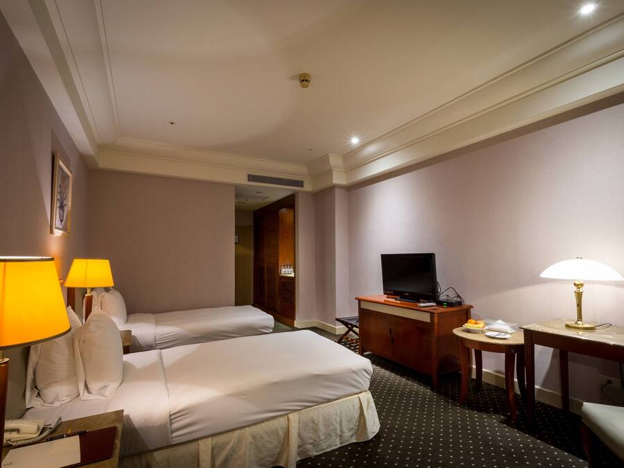 A hotel room with two beds, a desk with a TV, a desk lamp, a telephone, and a small table with a lamp.