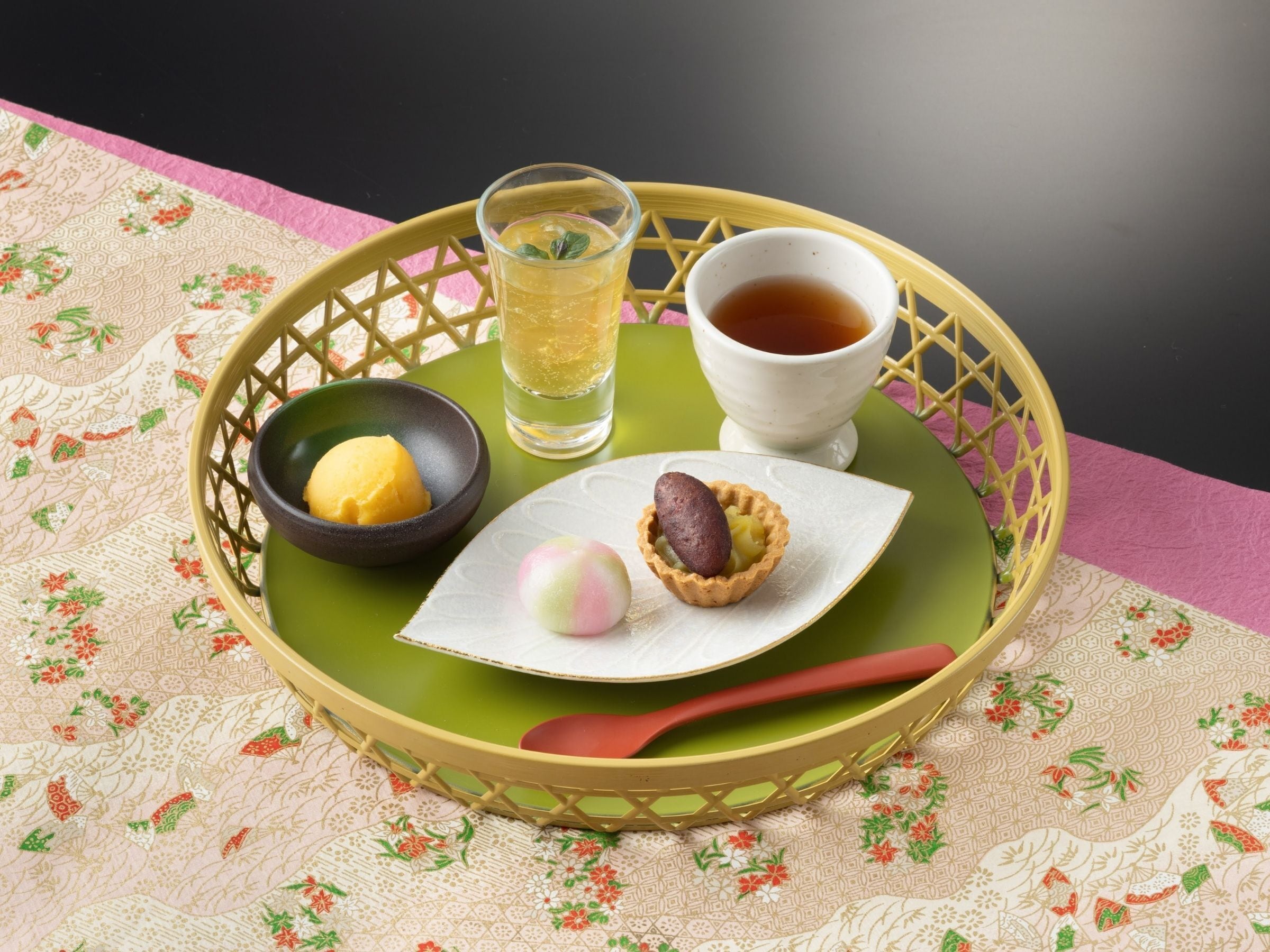 Una bandeja con una taza de té, un vaso de agua, un plato de comida y un pequeño postre sobre una mesa con mantel estampado de flores.