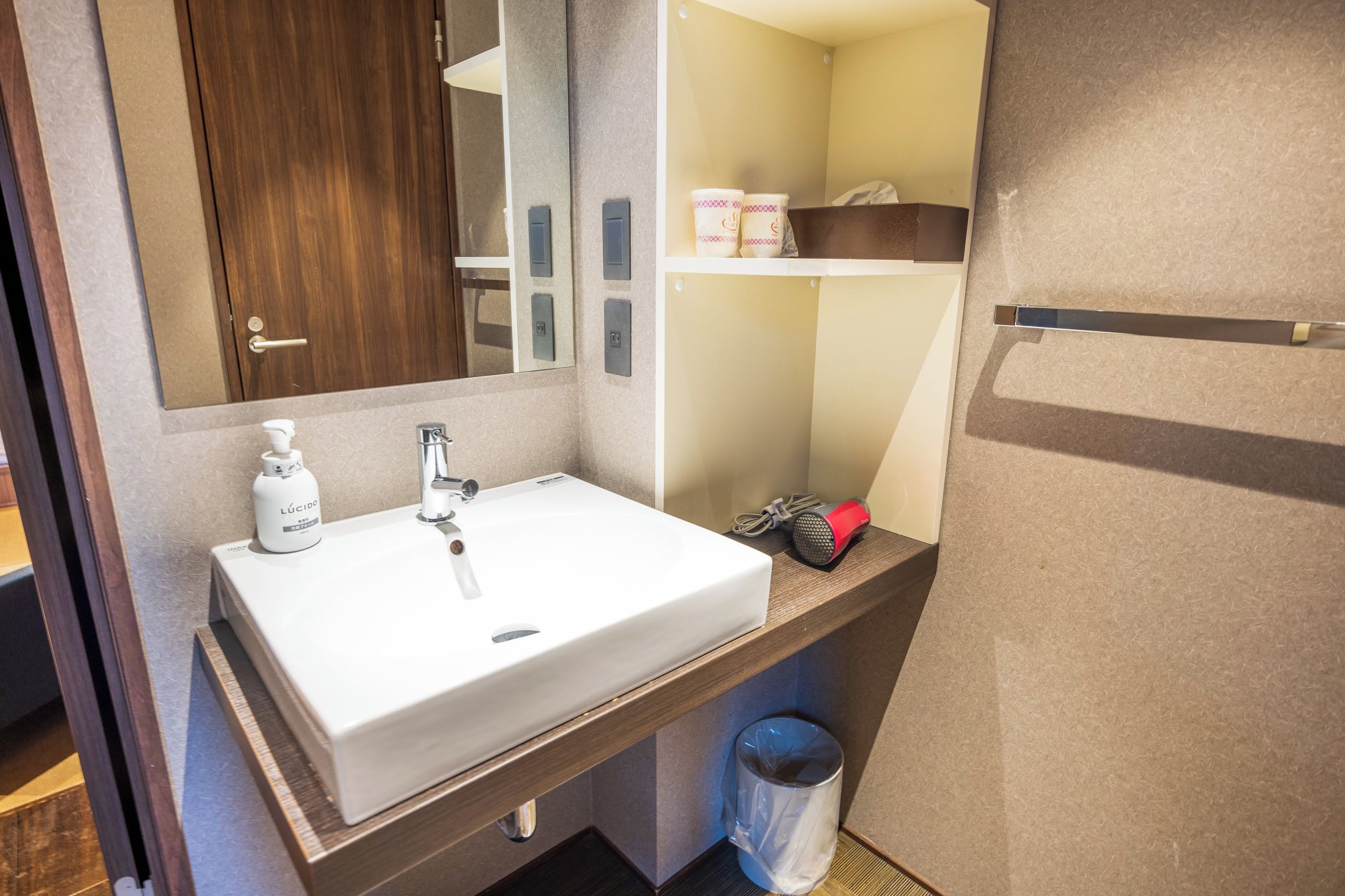 A bathroom with a white sink, a mirror, a towel dispenser, and a trash bin.