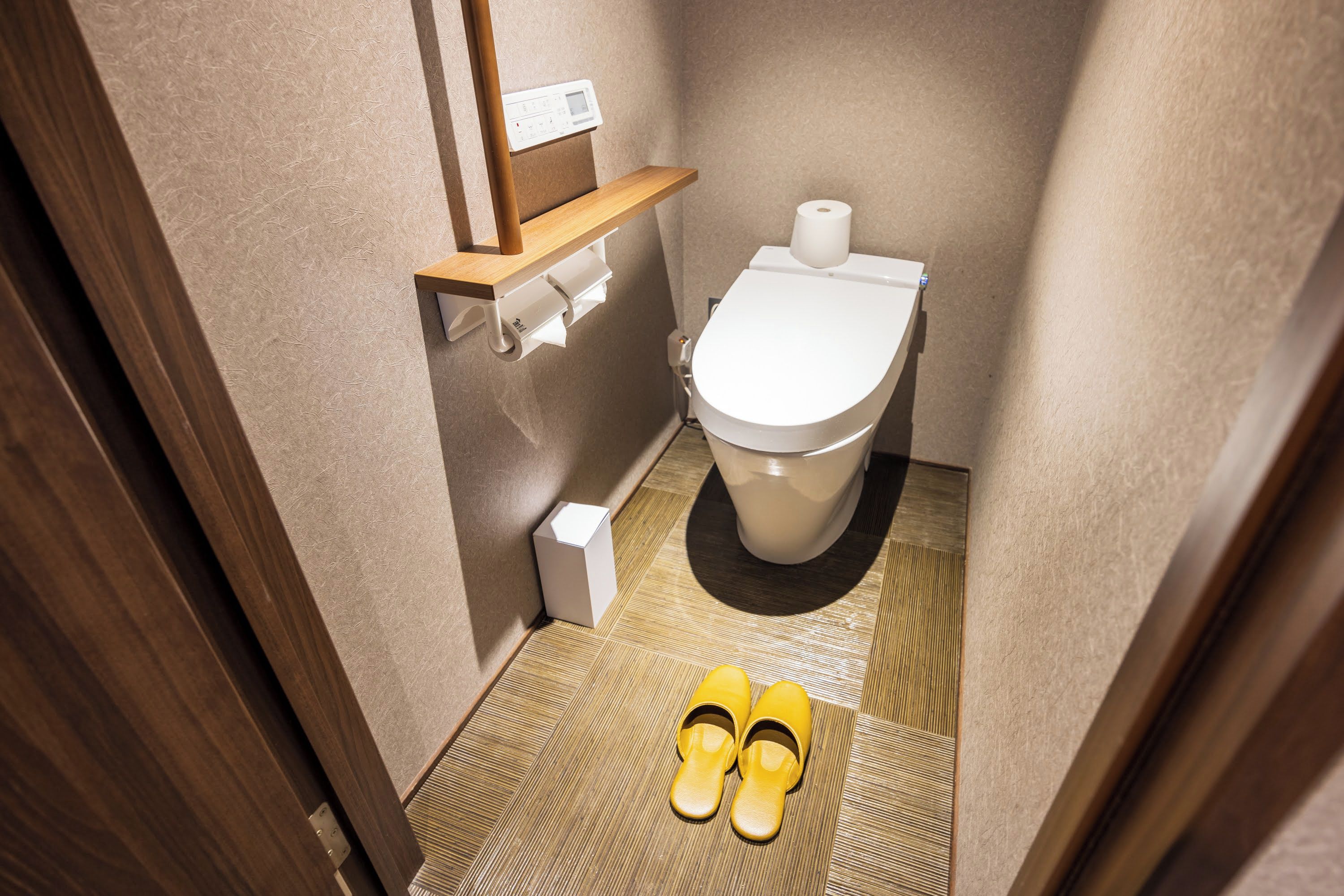 A small bathroom with a toilet, a shelf with a toilet paper holder, and a pair of yellow slippers on the floor.