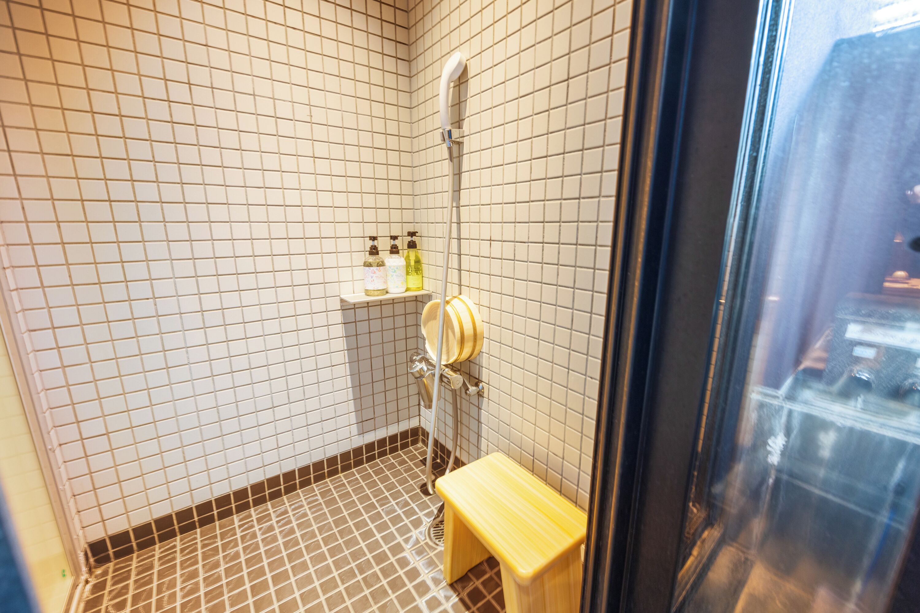 Ein kleines Badezimmer mit weißen und braunen Fliesen, einem hölzernen Hocker und einem Ablagefach mit Pflegeprodukten.