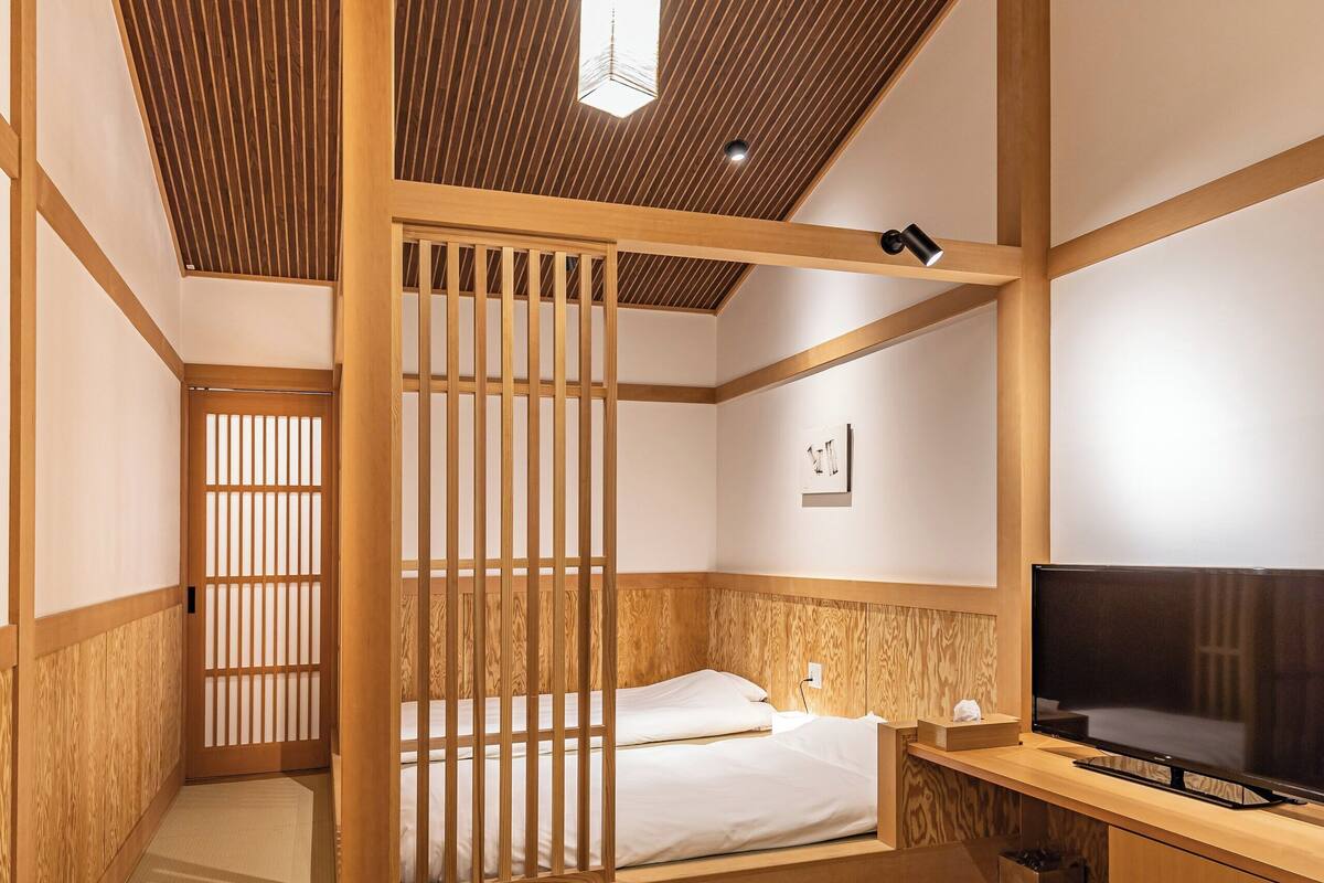 Ein traditioneller japanischer Raum mit Holzboden, einem Bett mit weißer Bettwäsche, einem Fernseher auf einem hölzernen Standfuß und einem Tatami-Boden.