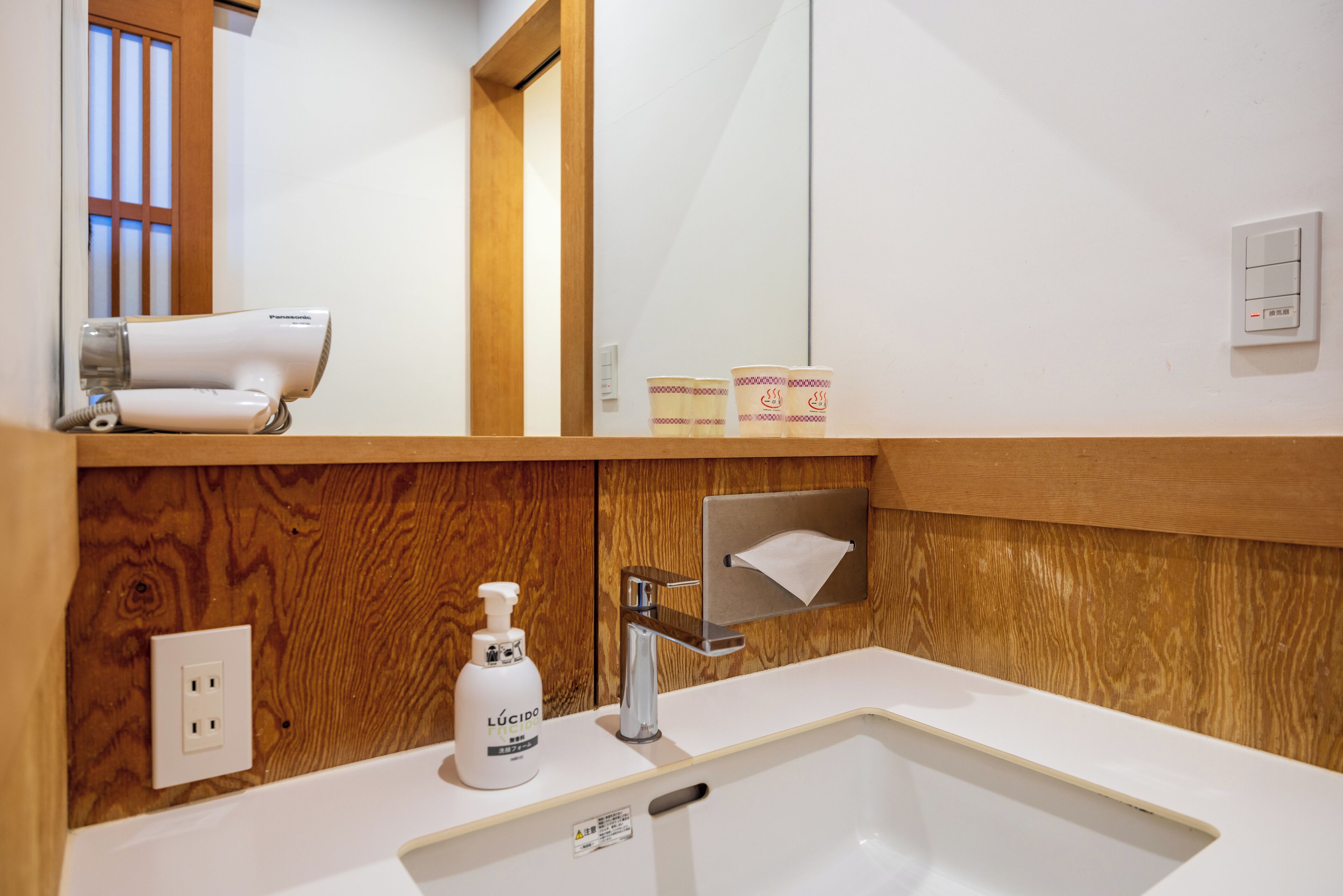 Un baño con un tocador de madera, un lavabo blanco, un espejo, un secador de pelo y una botella de jabón Lúcido.