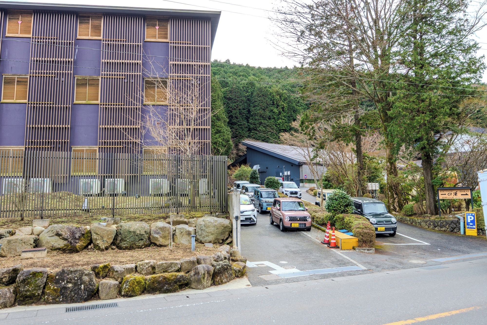 A parking area with several cars, a building with vertical wooden slats, and a stone wall with rocks.