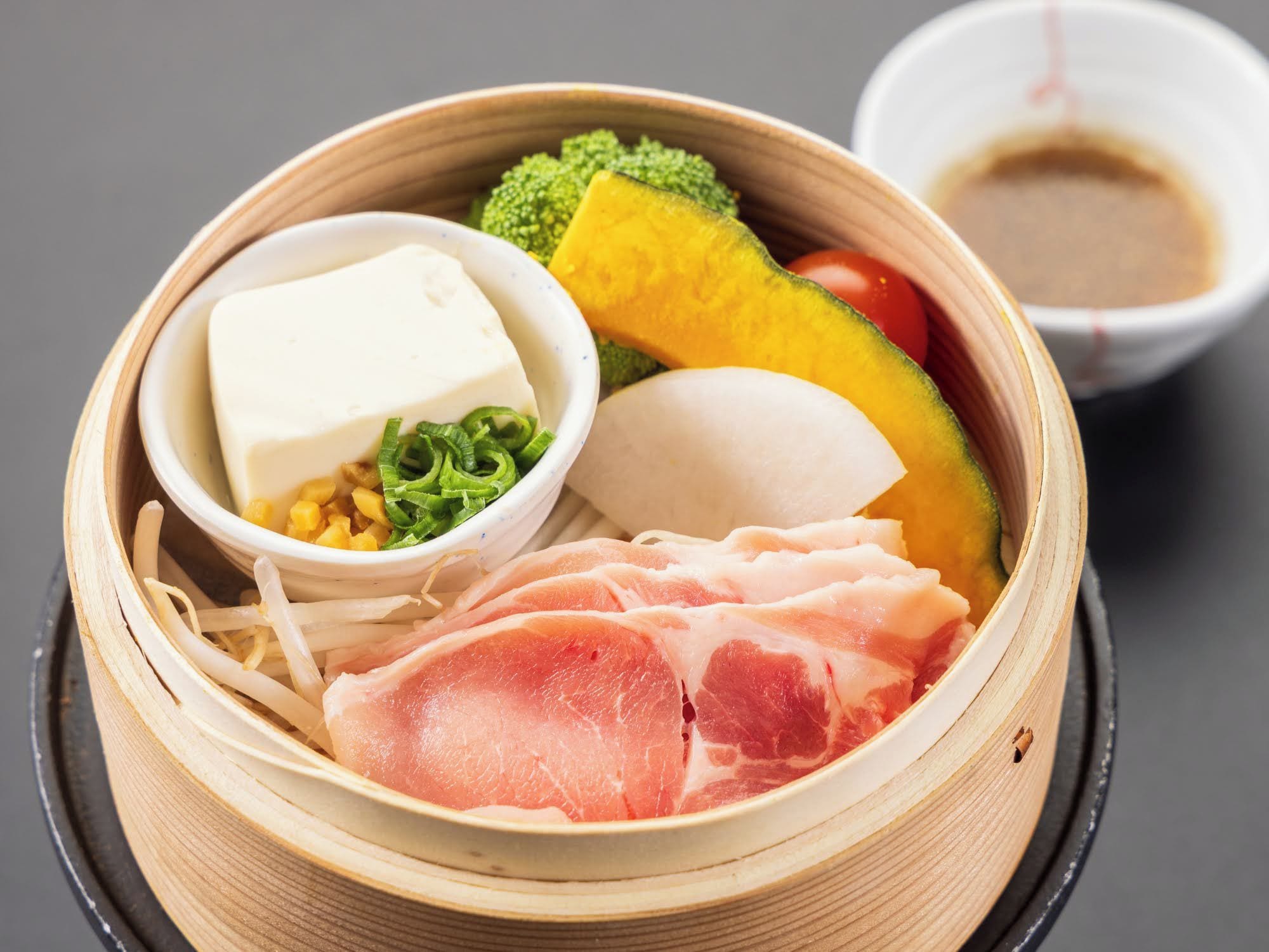 A bento box with various food items including meat, vegetables, and a small bowl of sauce.