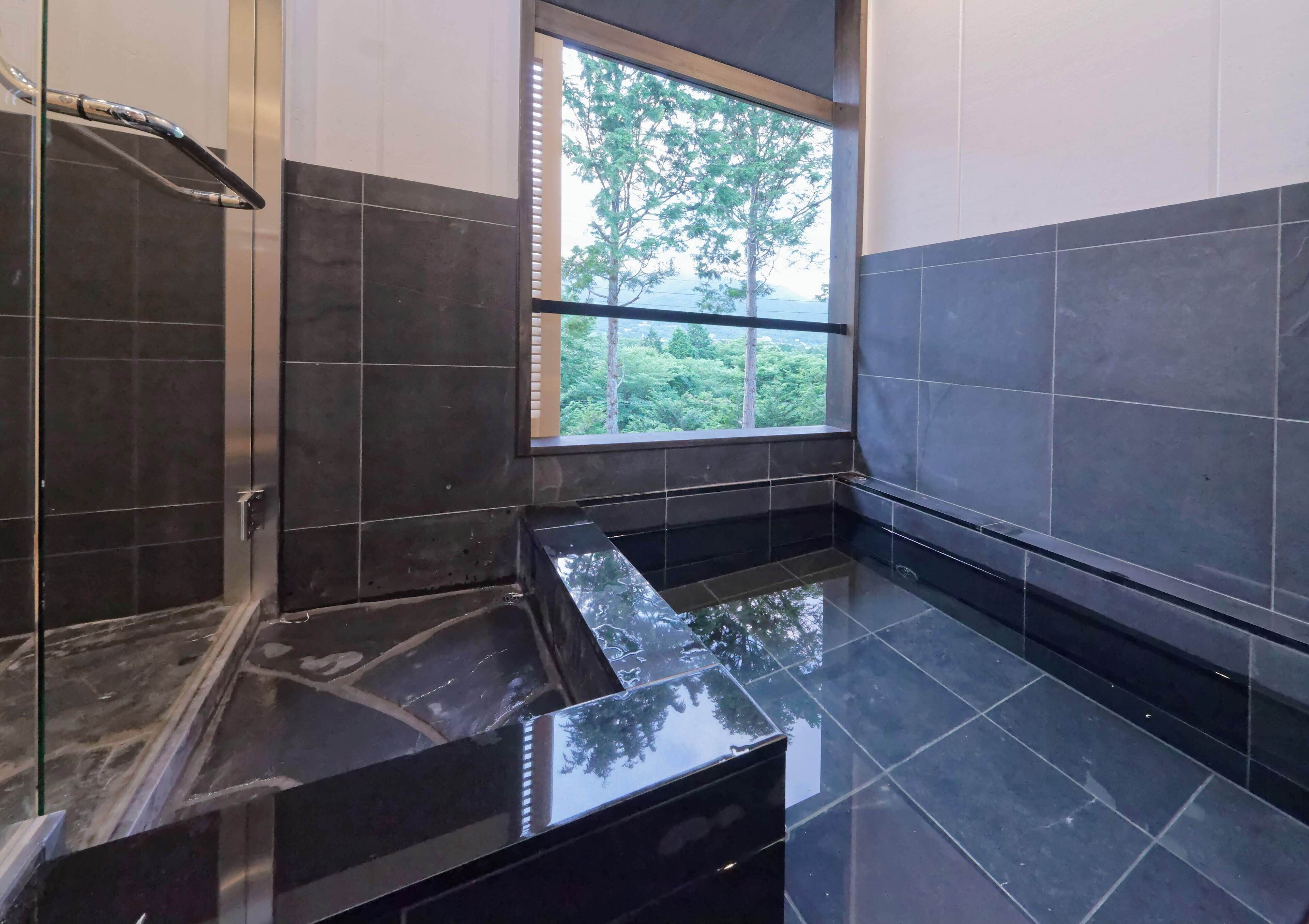 A modern bathroom with dark tiled walls, a glass shower enclosure, and a window with a view of trees.