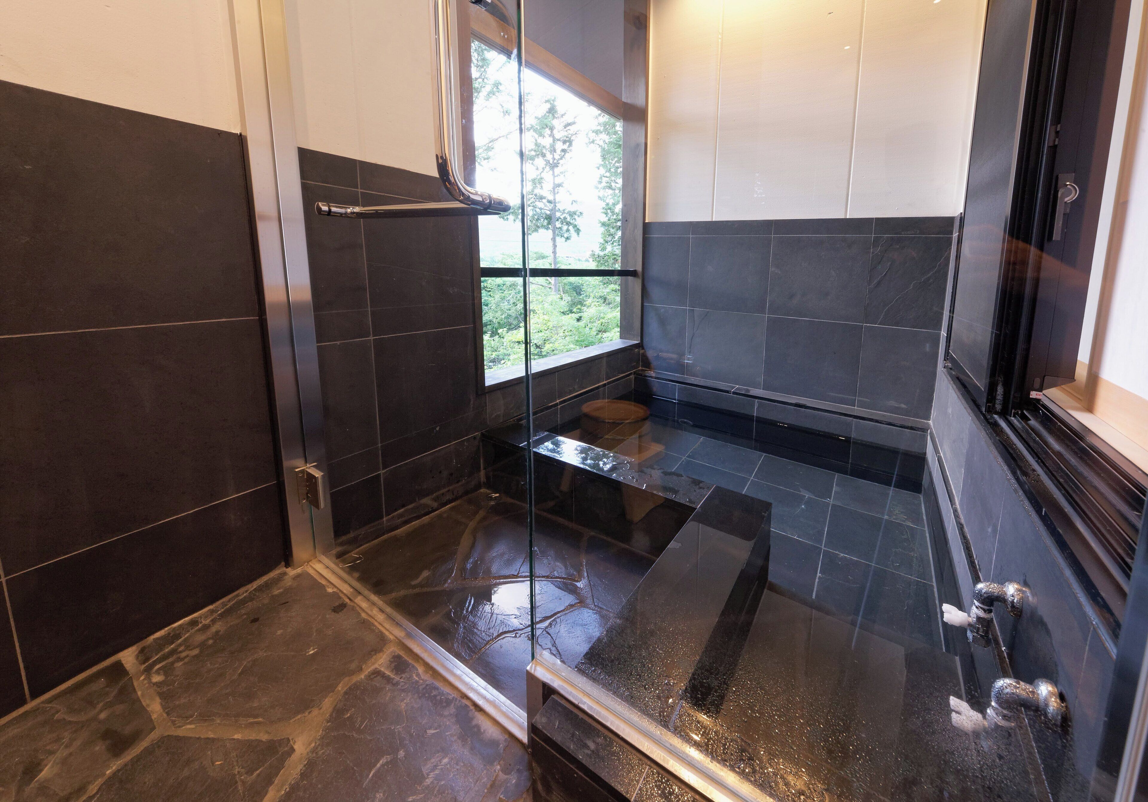 A modern bathroom with dark tiles, a glass shower enclosure, and a window providing a view of greenery.