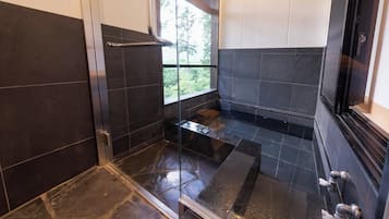 A modern bathroom with dark tiles, a glass shower enclosure, and a window providing a view of greenery.
