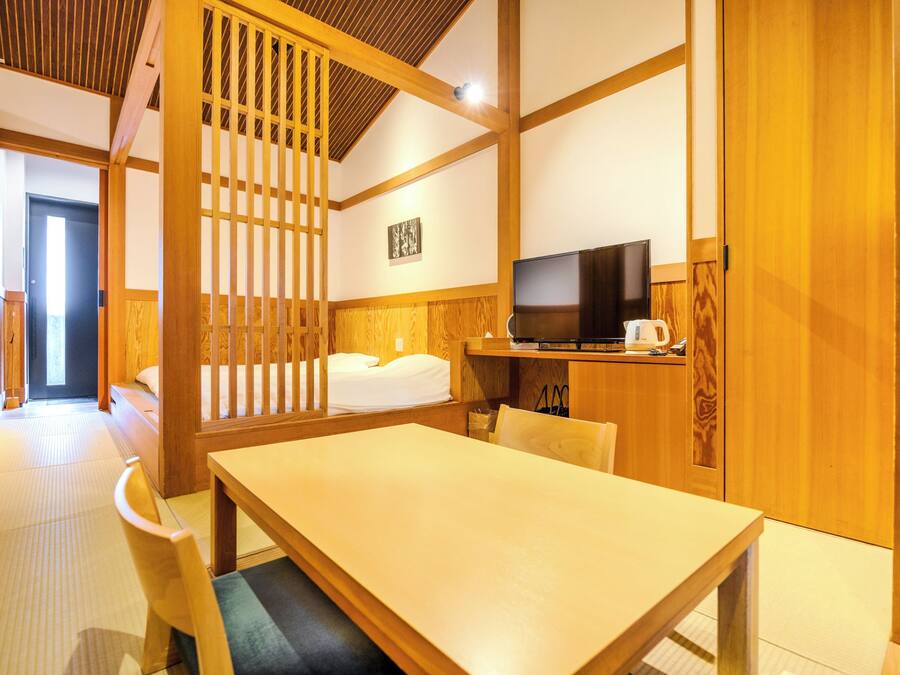 A traditional Japanese-style room with tatami flooring, a low table, a bed with white bedding, a television, and a wooden cabinet.