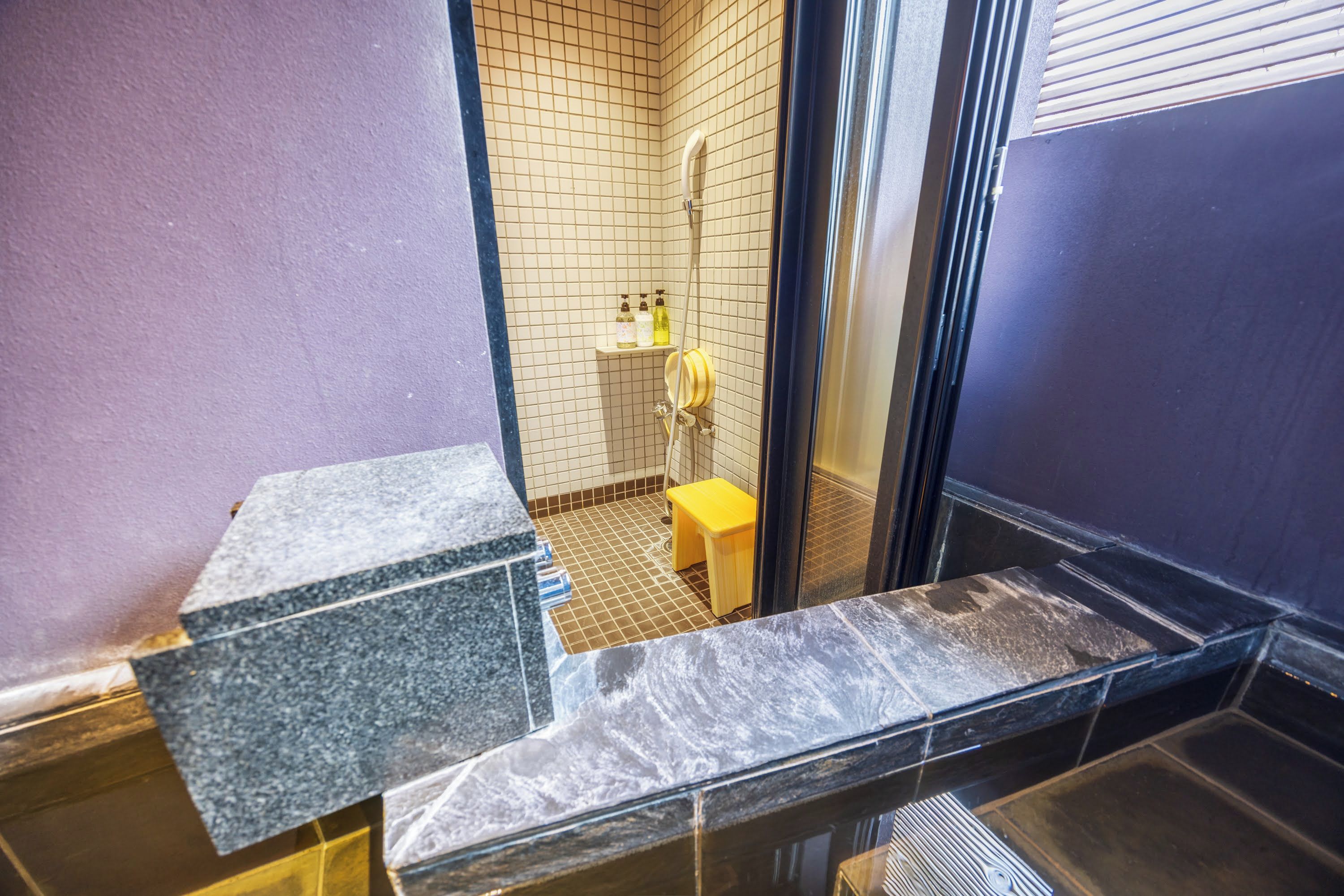 Un baño con piso de baldosas, zona de ducha y un taburete pequeño amarillo.
