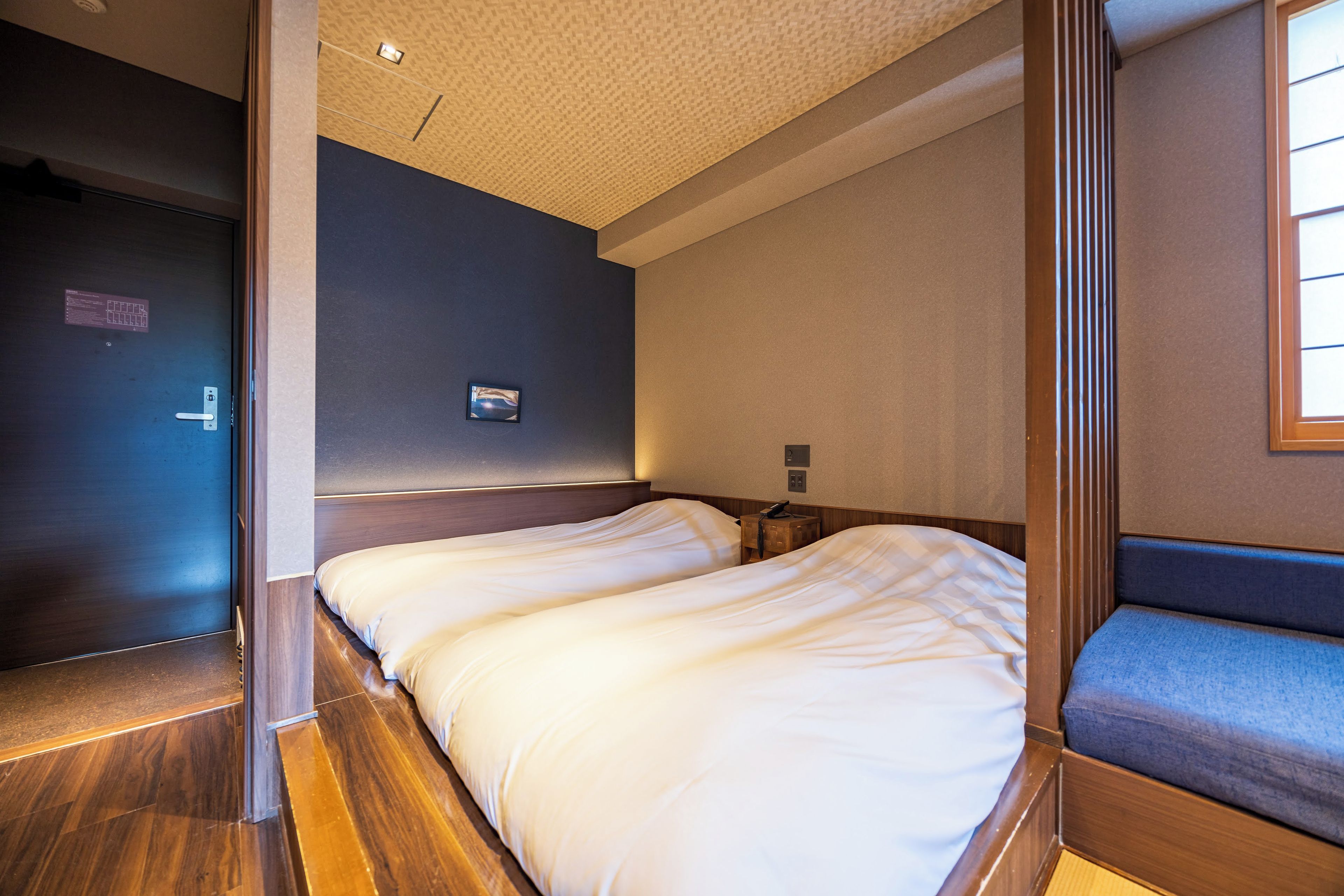 Une chambre d’hôtel avec deux lits, un plancher en bois, un mur sombre, un petit tableau et une porte.