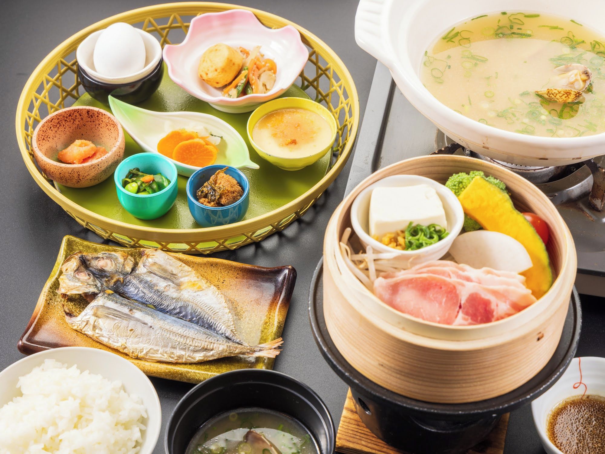 A variety of dishes including fish, rice, and soup, served on a table with a basket and a steamer.