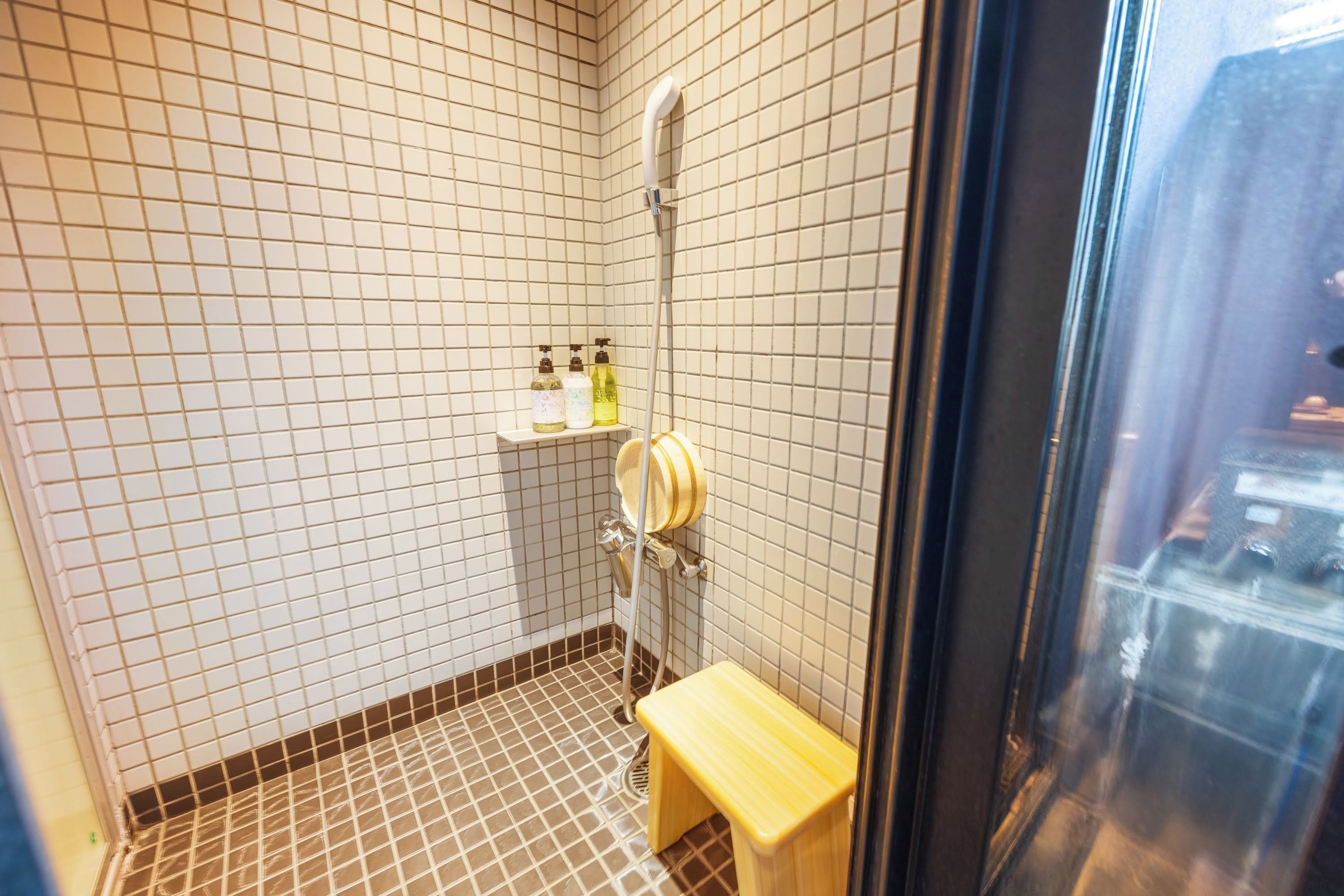 A small bathroom with white and brown tiled walls, a shower, and a wooden bench.