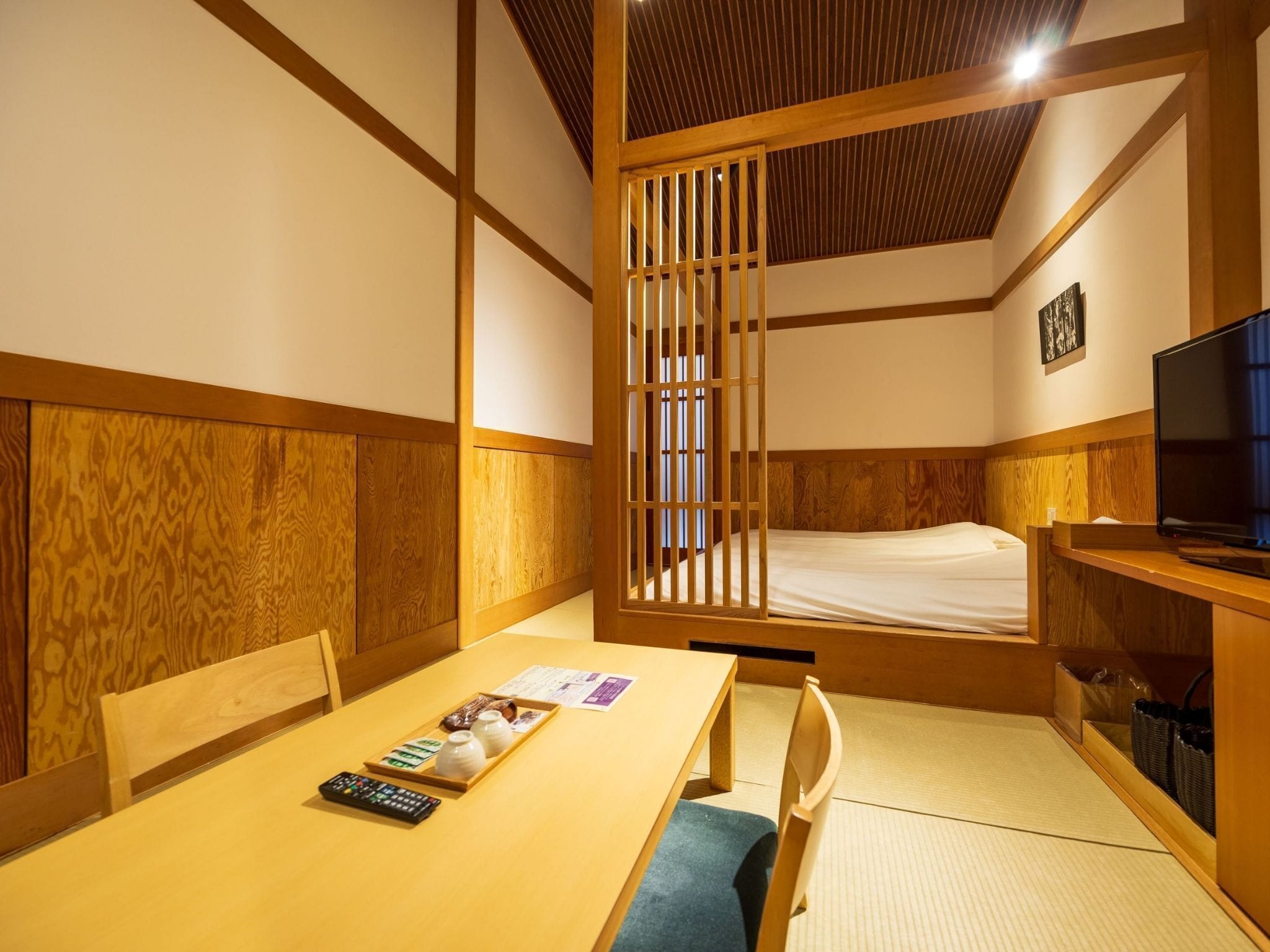 A traditional Japanese-style room with tatami flooring, a low wooden bed, a television, and a table with a tray of snacks and a remote control.