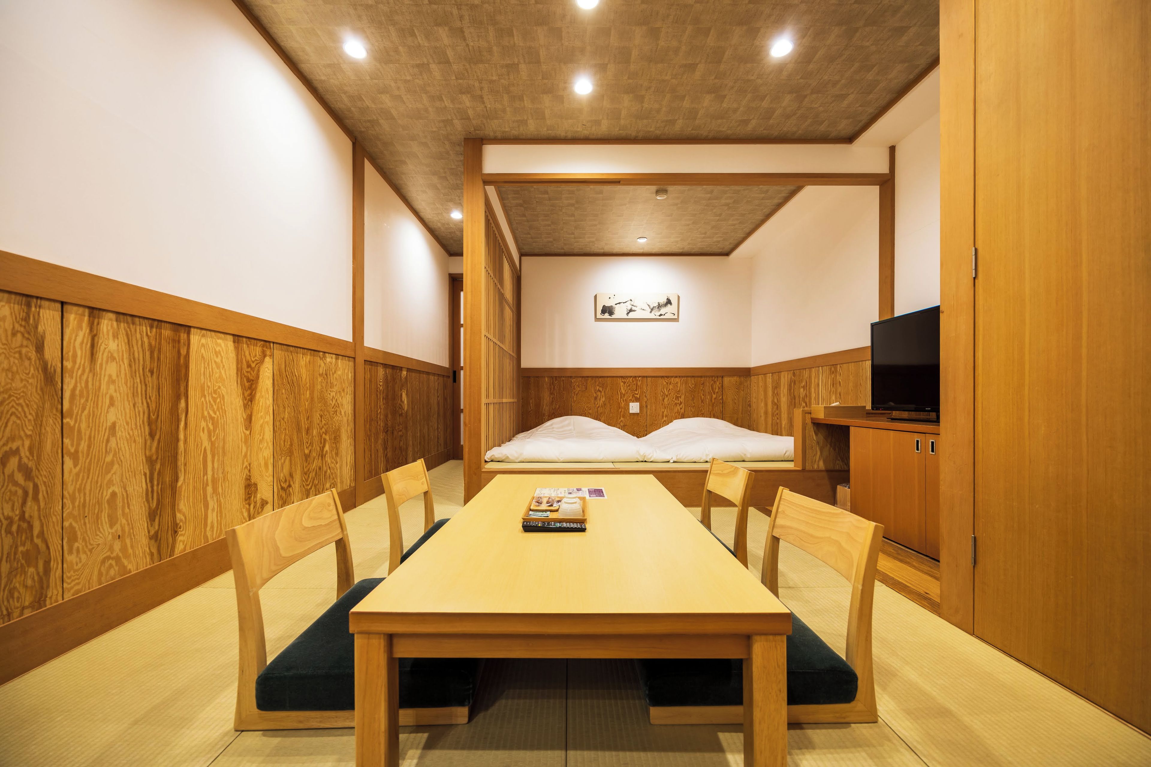 A room with a wooden table and chairs, a bed with white bedding, a television on a wooden cabinet, and a wall-mounted artwork.
