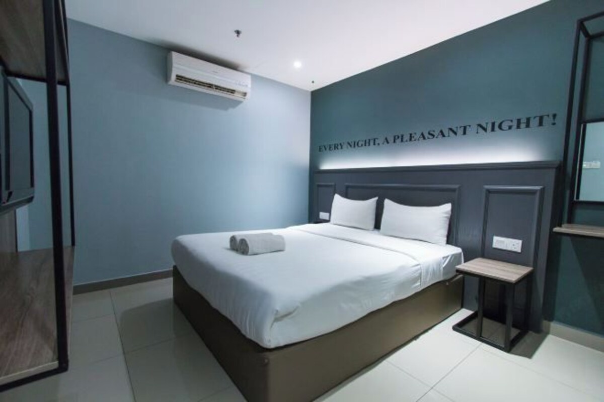 Une chambre d’hôtel avec un lit, une petite table de chevet, un appareil de climatisation et un mur sur lequel est inscrit le texte : « TOUTES LES NUITS, UNE NUIT AGRÉABLE ! ».