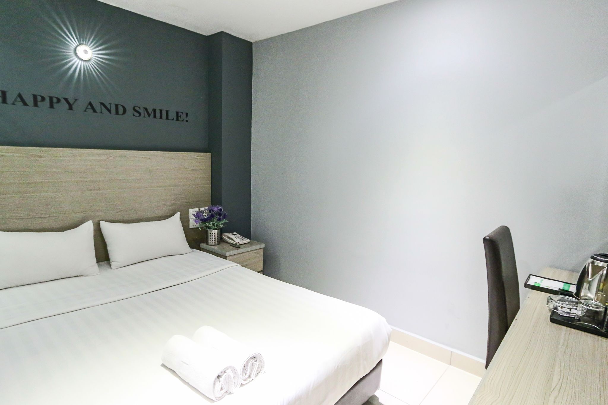 A hotel room with a bed, a nightstand with a phone and a vase of flowers, a desk with a coffee maker, and a wall with a 'Happy and Smile!' sign.